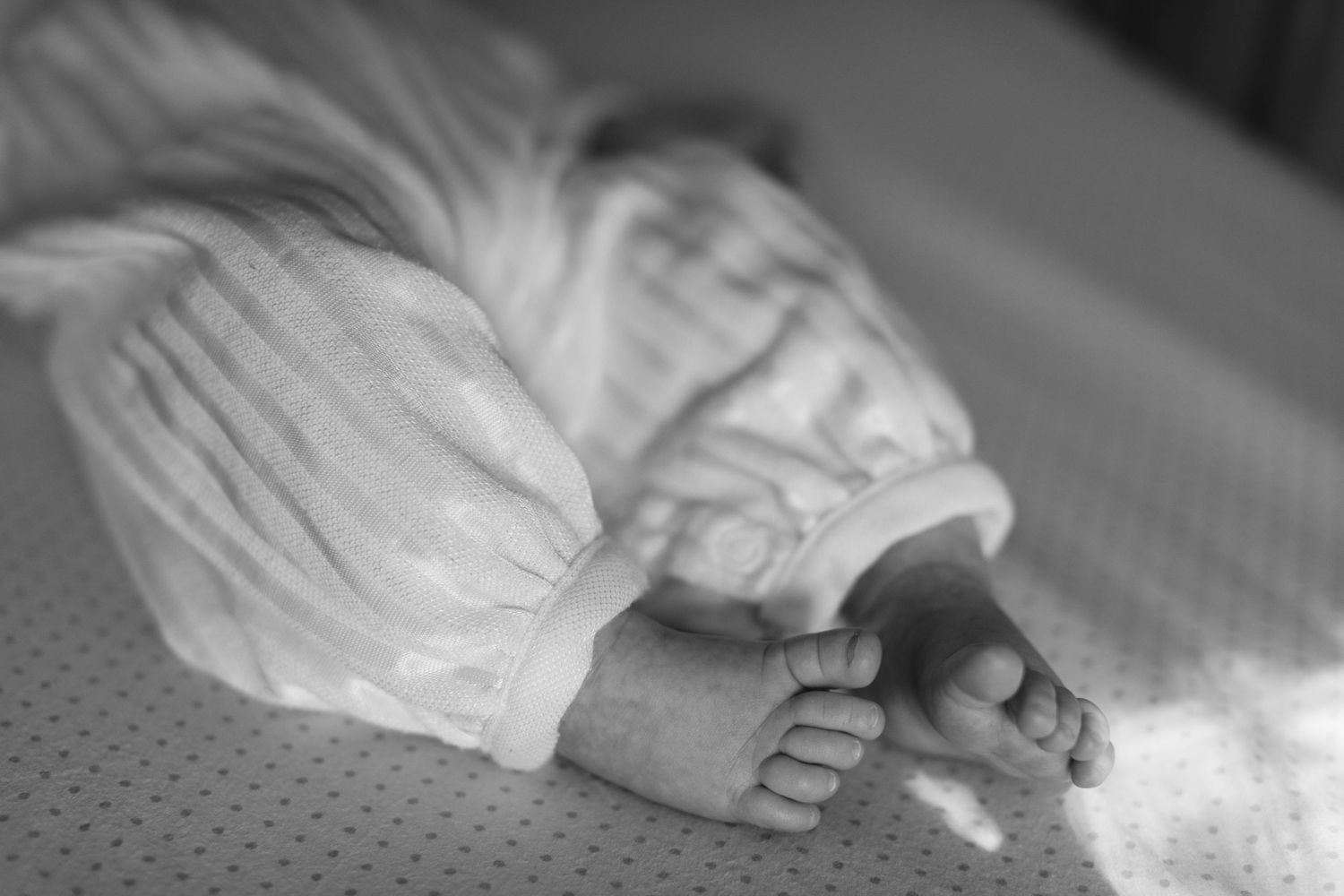 Black and white close-up of newborn baby tiny feet in white outfit on soft mattress.