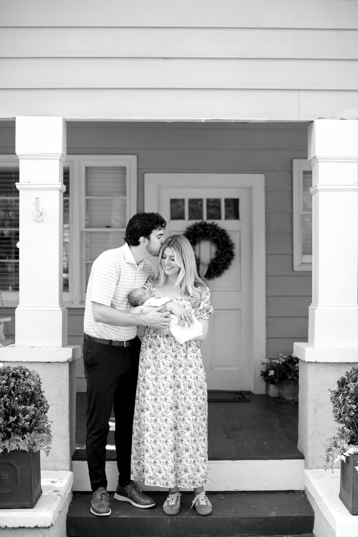 Black and white photo of couple on porch steps, man kissing woman's head as she holds newborn.