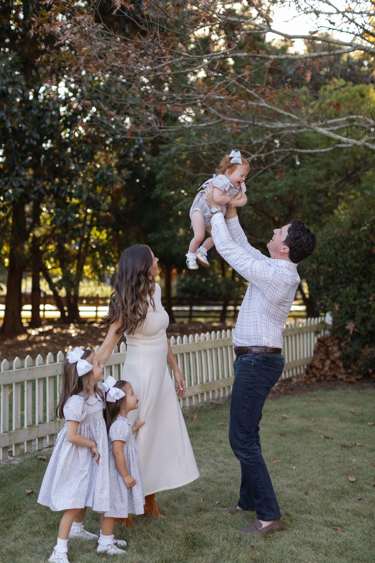 Family members in coordinating white outfits play together on a grassy lawn near a picket fence.