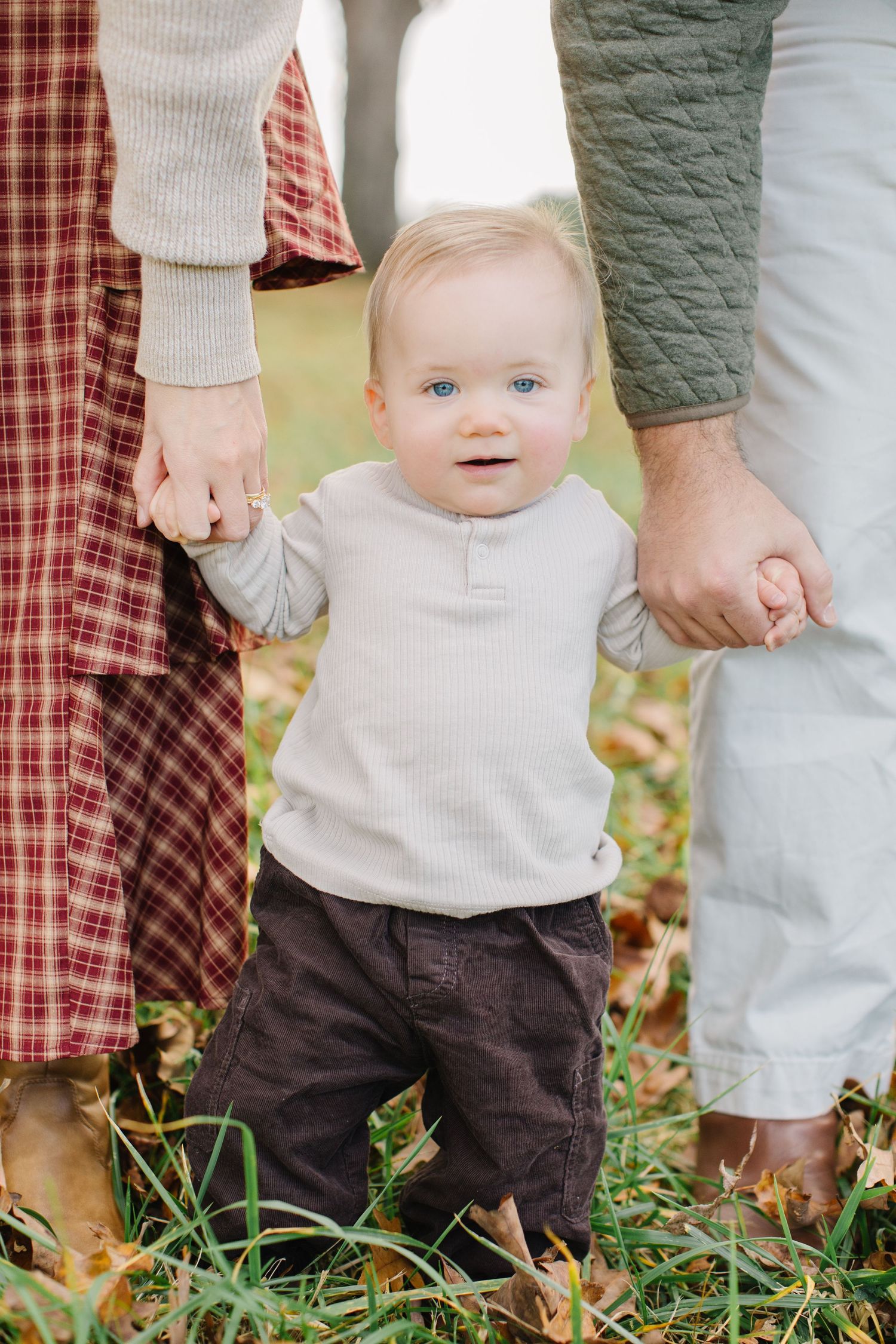 Small child stands between two adults in casual fall attire on grass with autumn leaves.