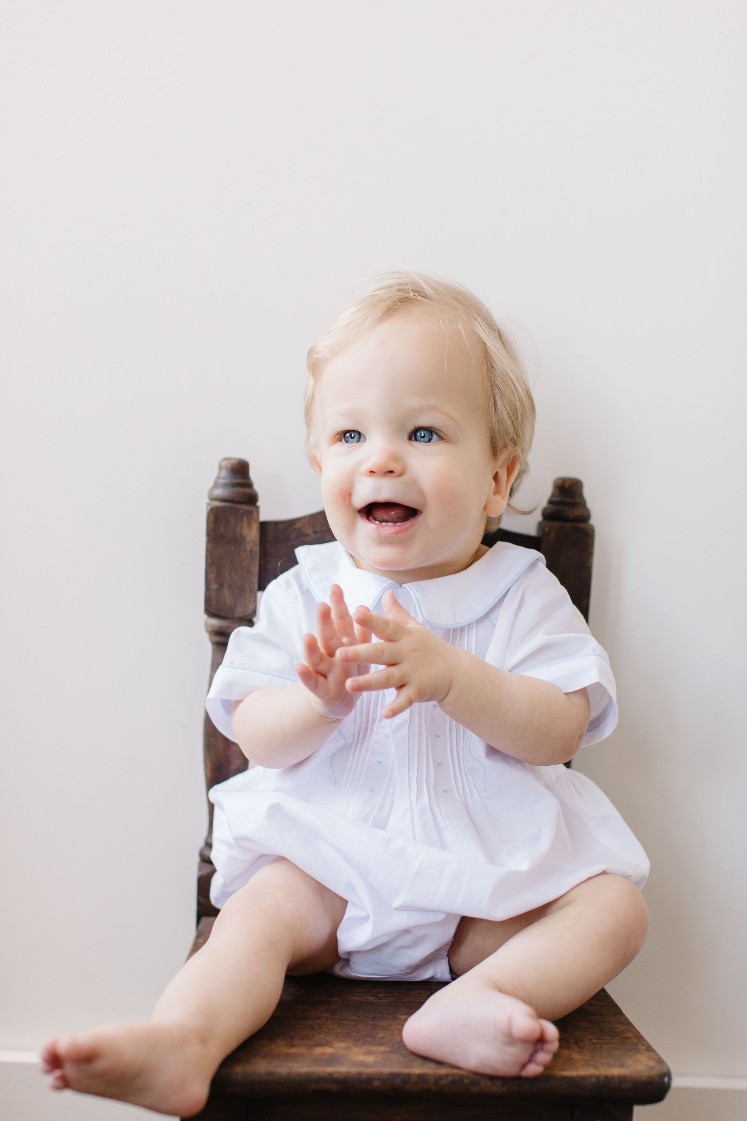 Happy baby in white outfit clapping and laughing while seated on brown wooden chair.