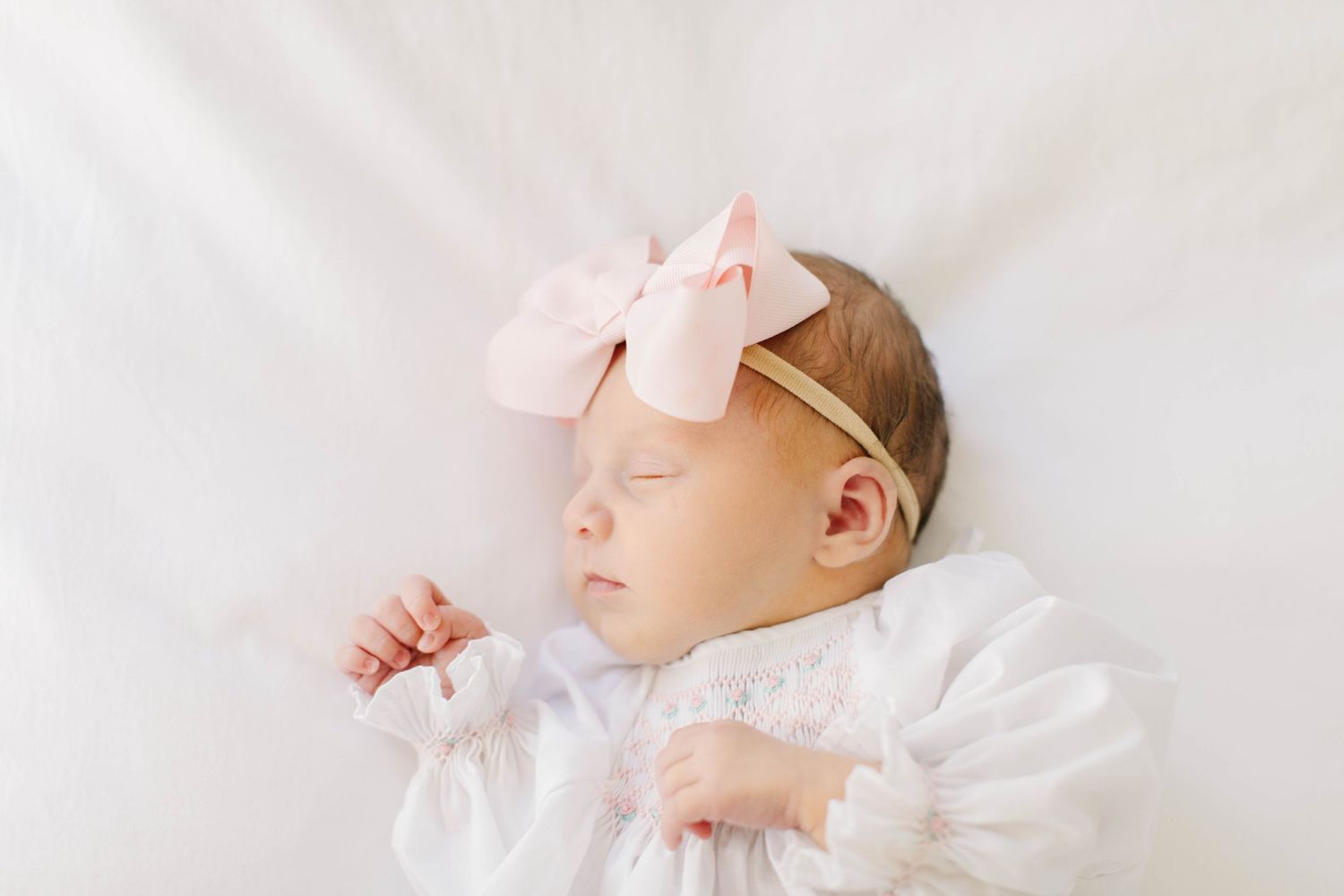 Sleeping newborn in white dress with pink bow accessory.