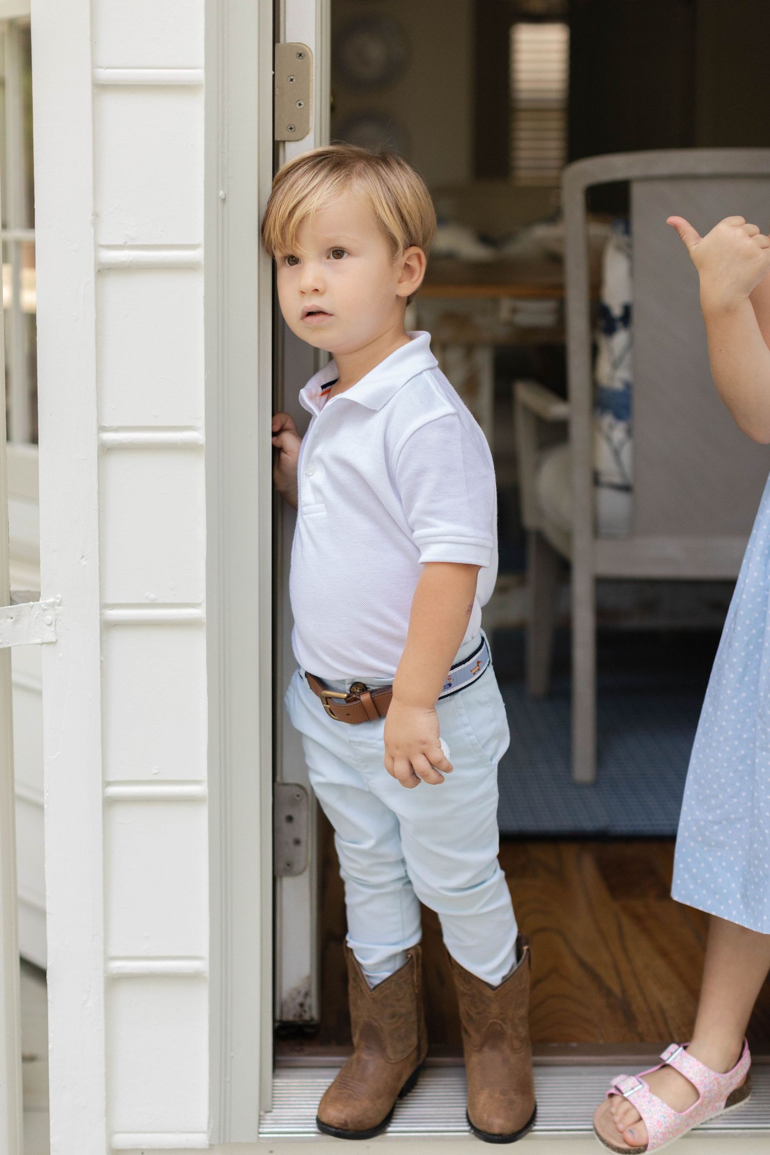Toddler in white shirt and blue pants wearing brown cowboy boots stands between white porch pillars.