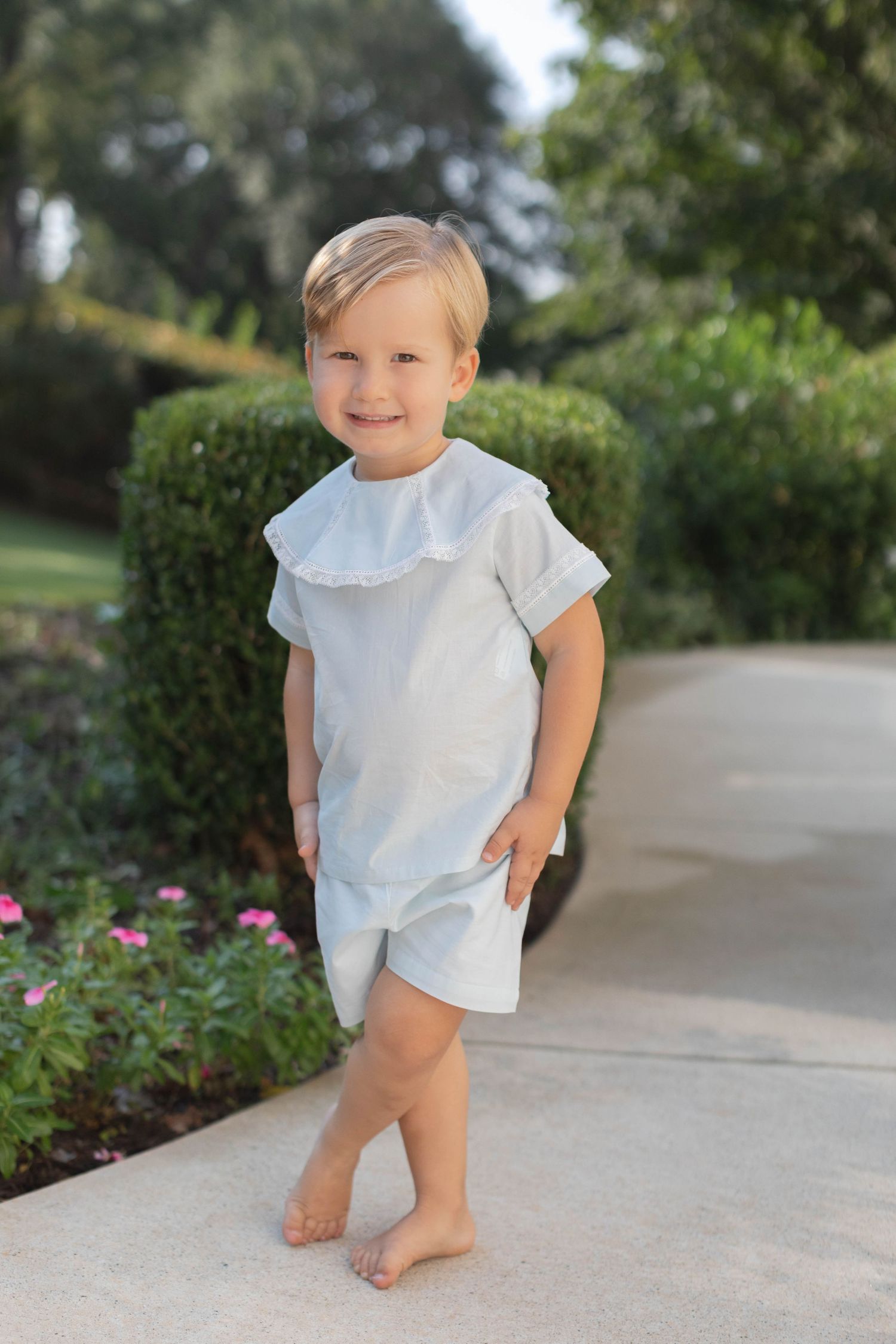 Small child in a light colored ruffle dress plays outdoors near pink flowers and green bushes in a garden setting.