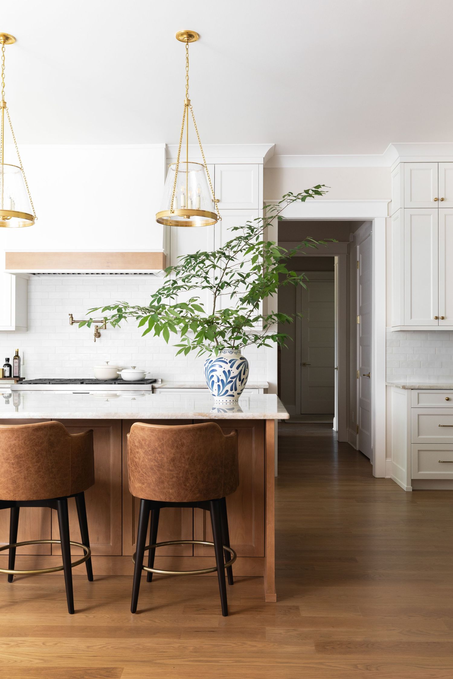 Modern white kitchen with wood island, leather barstools, brass pendant lights, and blue-white vase with greenery.