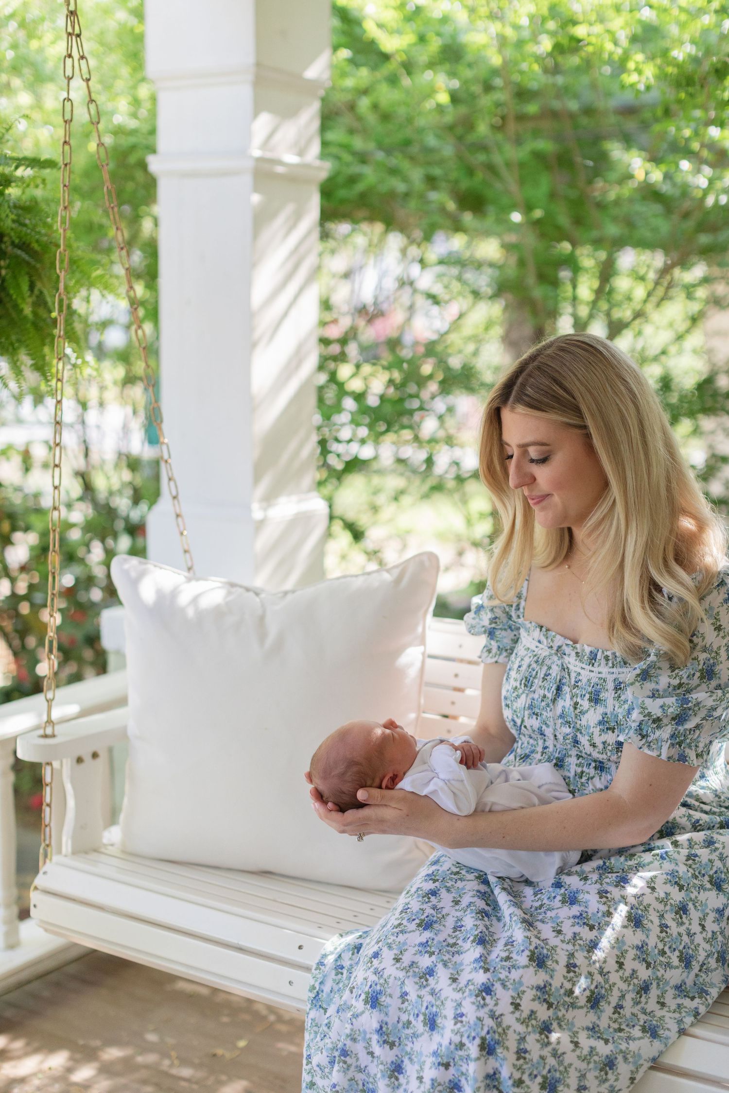 Blonde mother in floral dress holding newborn baby on white porch swing surrounded by lush greenery.