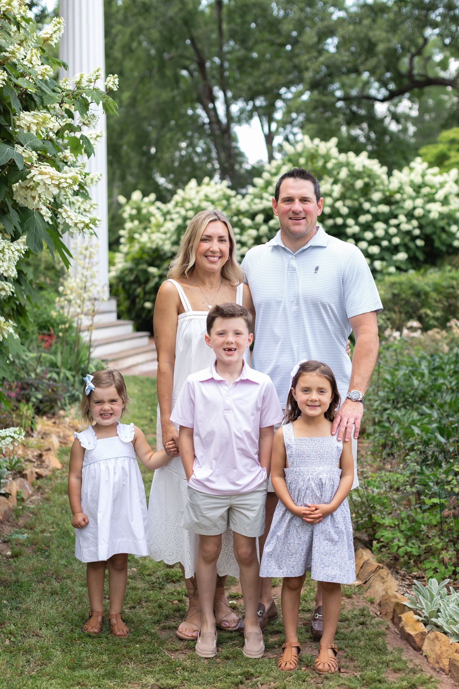 A happy family of five poses in a lush garden, parents with three young children dressed in pastel summer outfits.