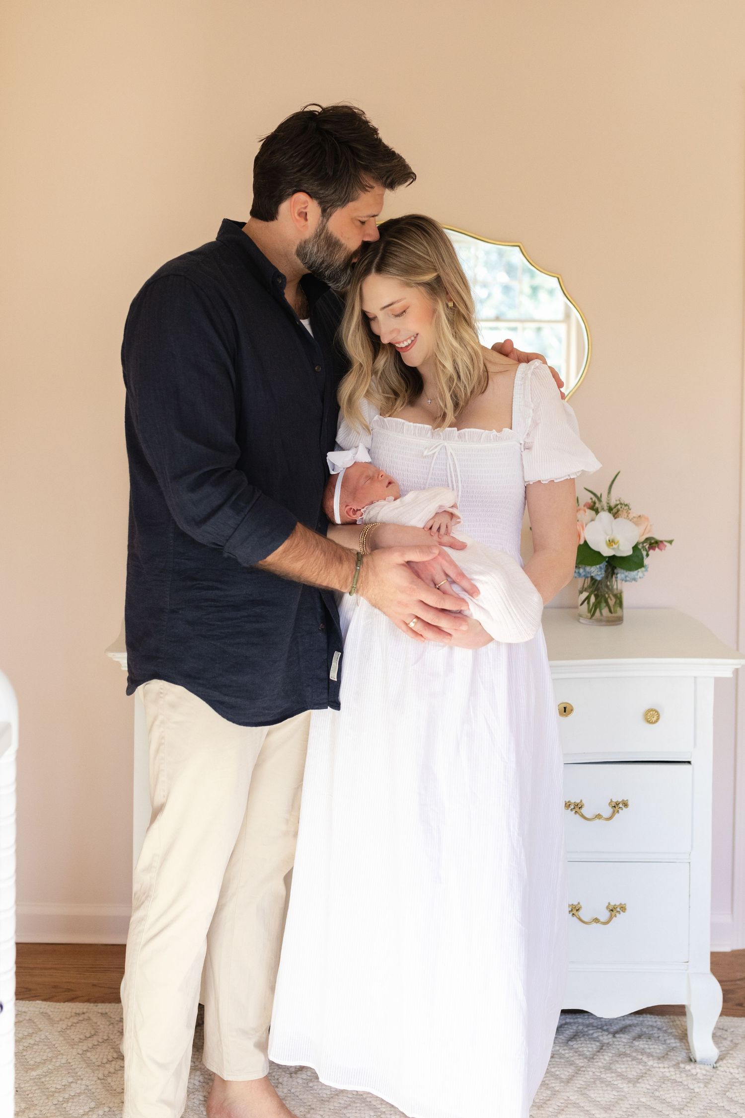 Parents in coordinated outfits share an intimate moment with their newborn in a nursery setting.
