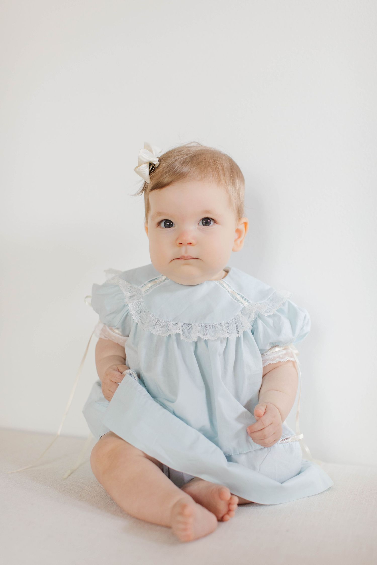 Baby sits in antique-style light blue dress with ruffled collar and bow accessory against white background.