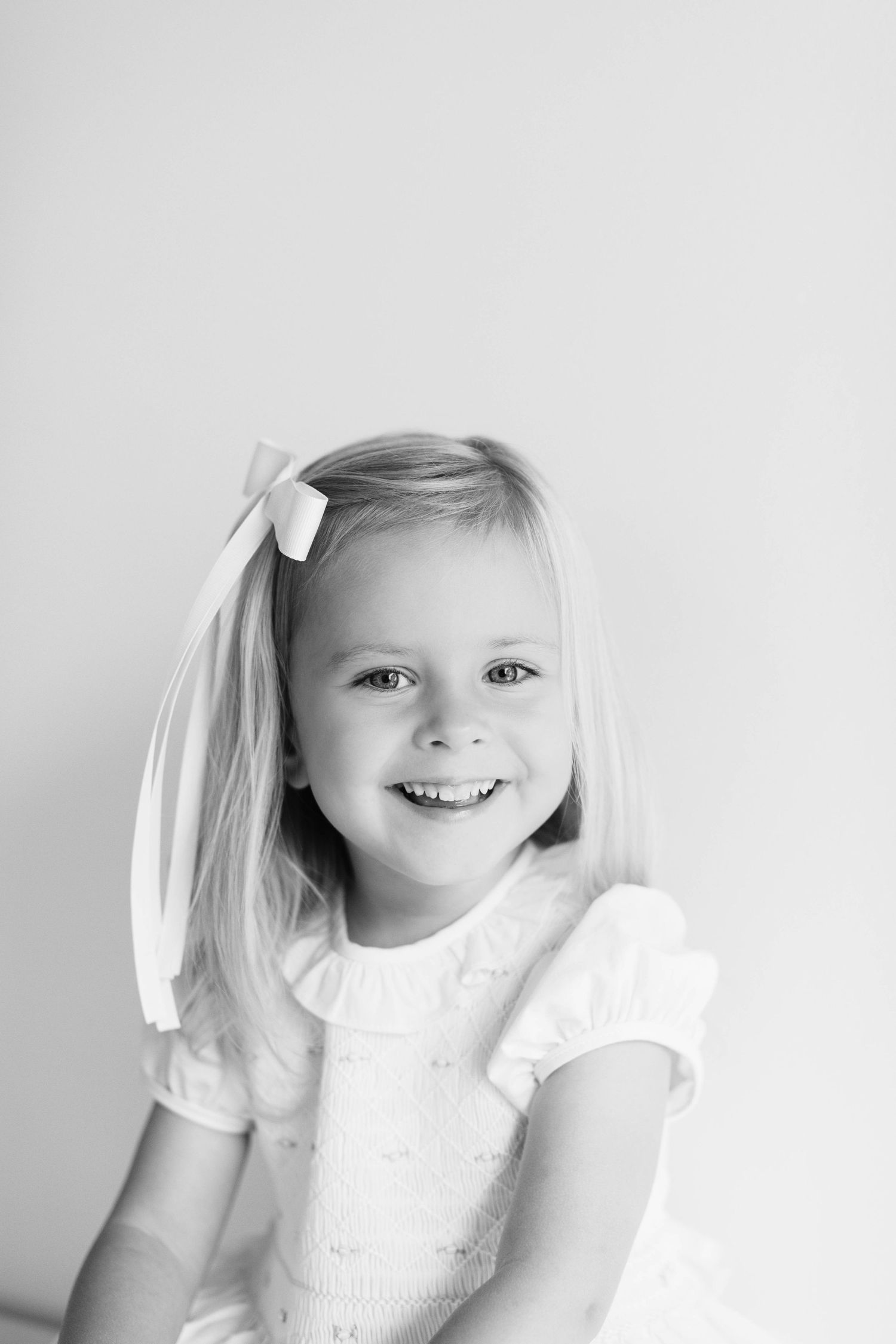 Black and white portrait of a young child wearing a white dress and hair ribbon smiling joyfully.