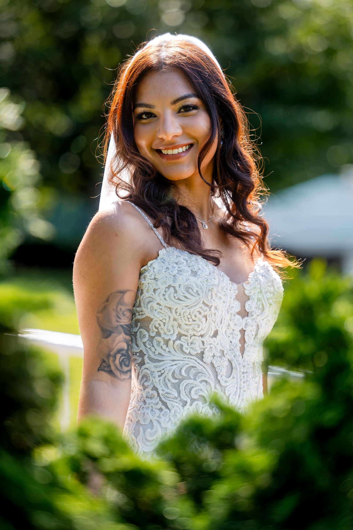 Person in white lace wedding dress smiling outdoors with natural greenery in background.