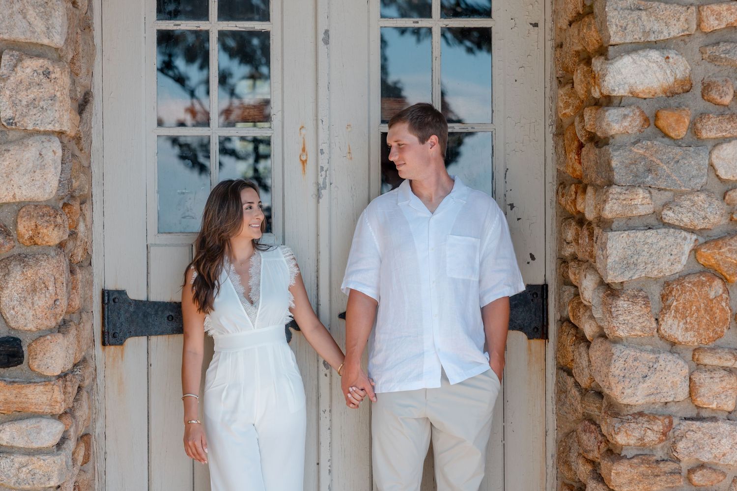 Couple laughs together against rustic stone wall with white trim windows.