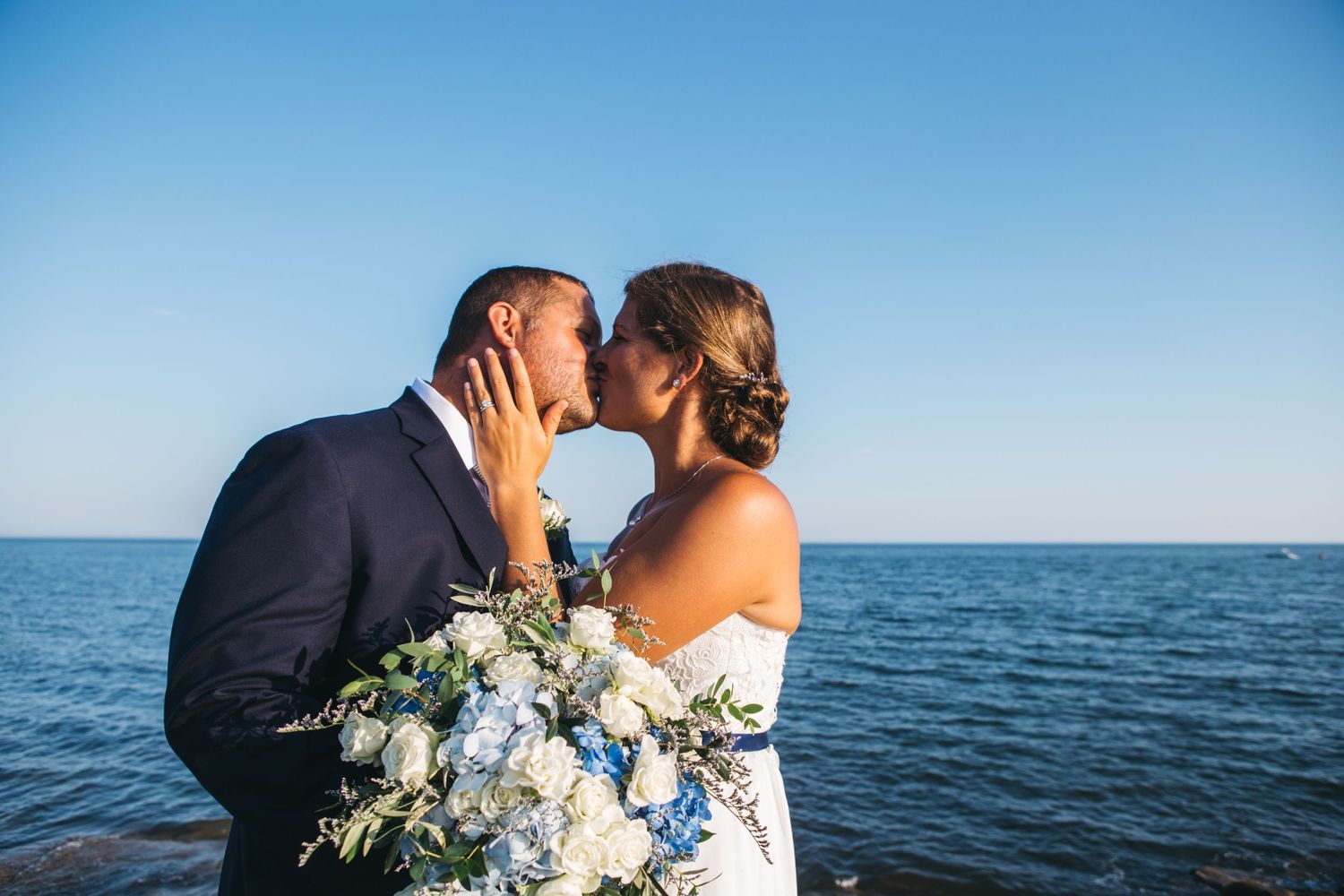 Romantic seaside wedding photoshoot with couple against blue ocean backdrop.