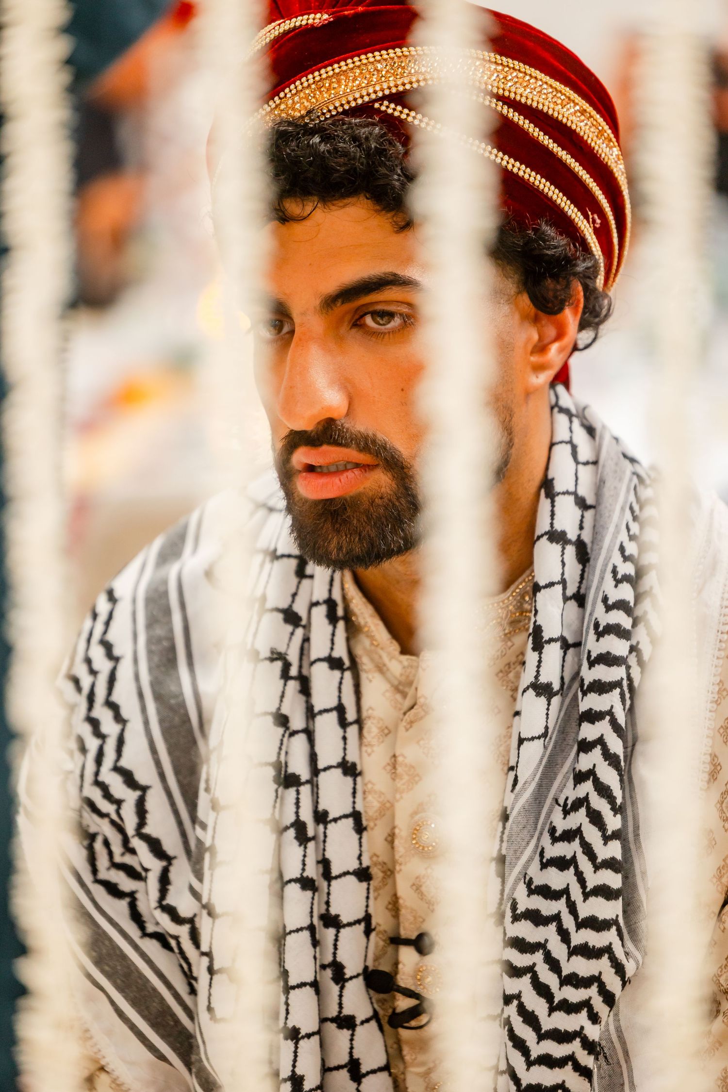 A groom wearing a red velvet turban and black and white patterned scarf is photographed through decorative elements.