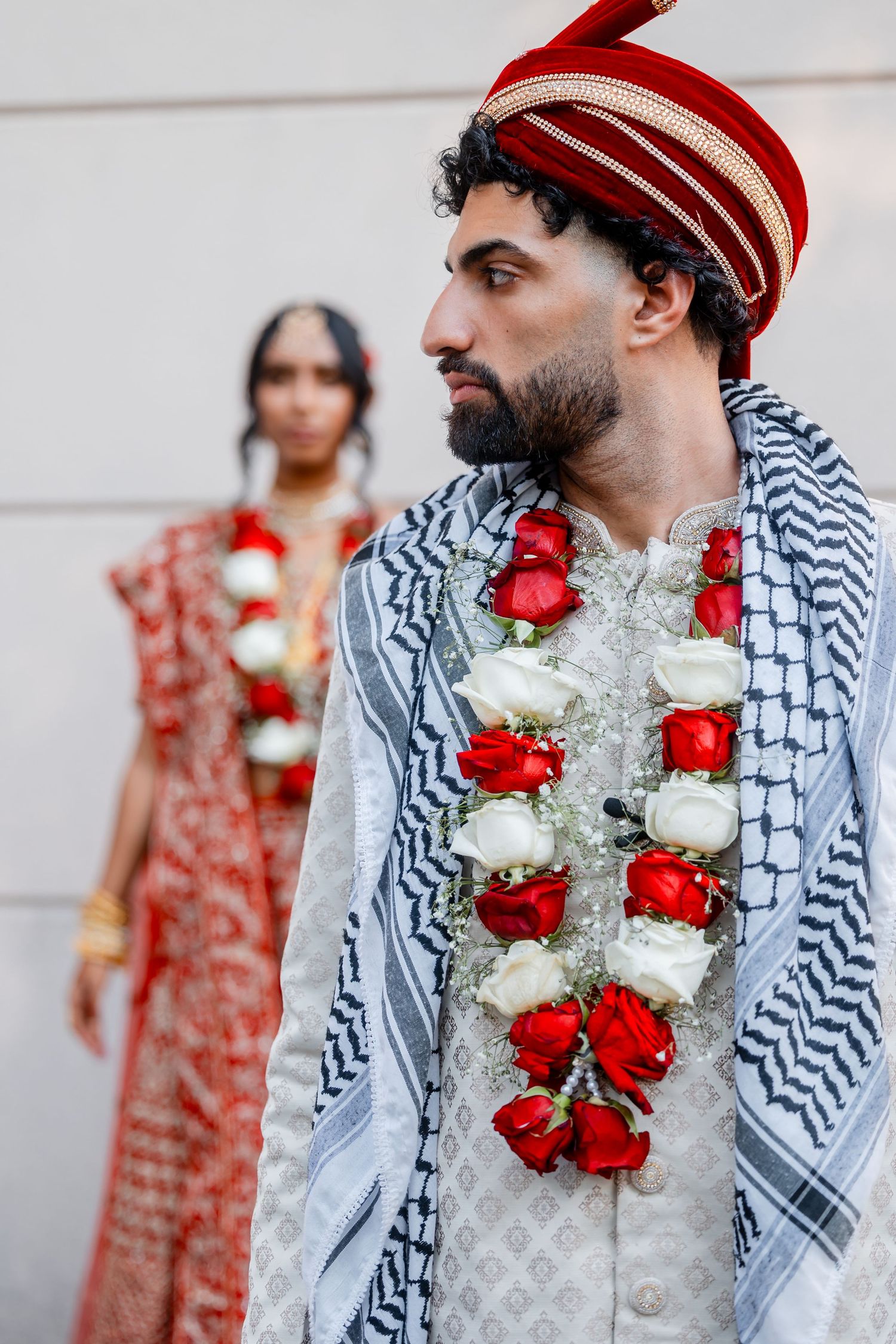 A groom in traditional attire with a red and white floral garland and striped headpiece stands near a wall.