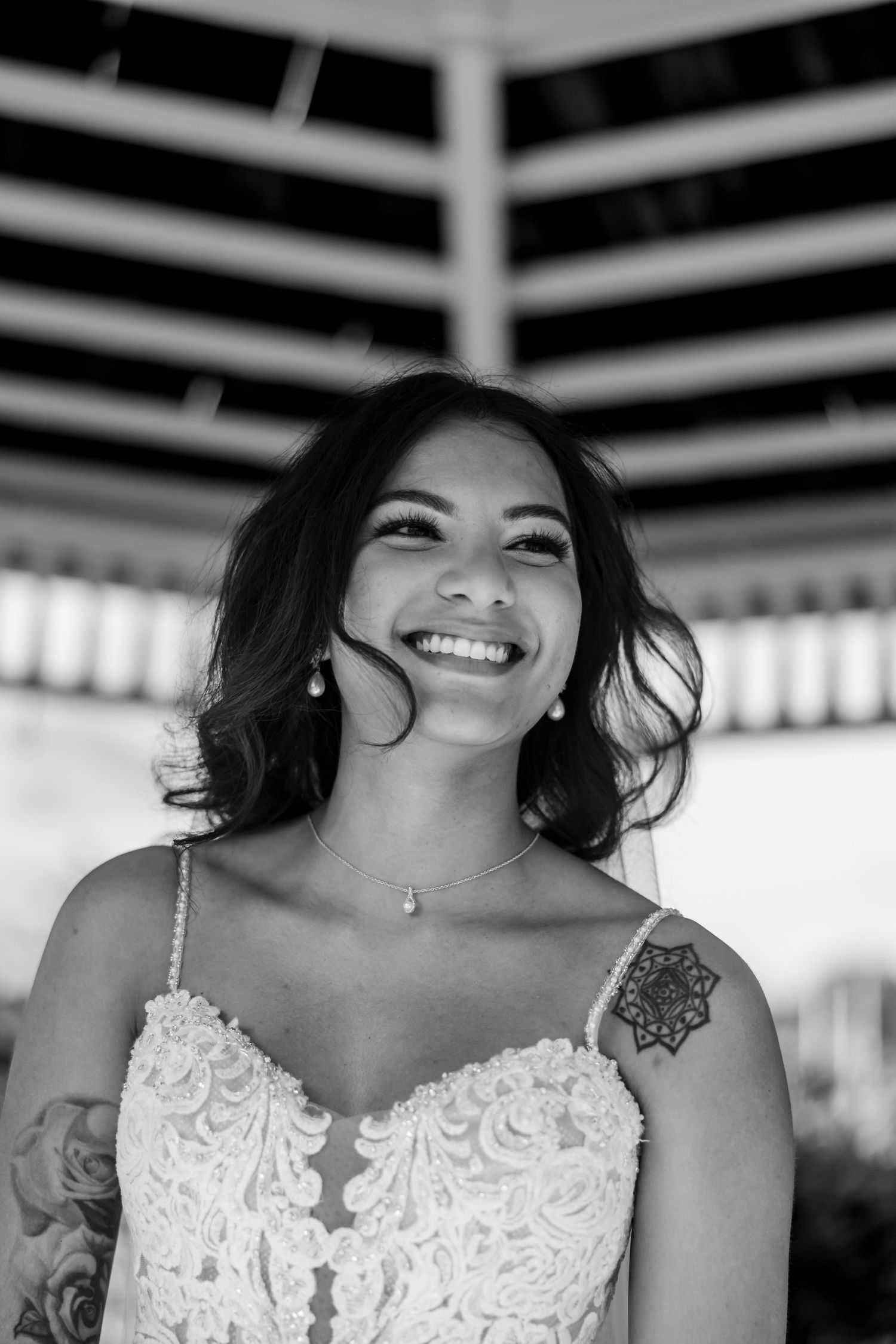 A bride wearing a white lace dress with delicate straps and shoulder tattoo poses under a covered area.