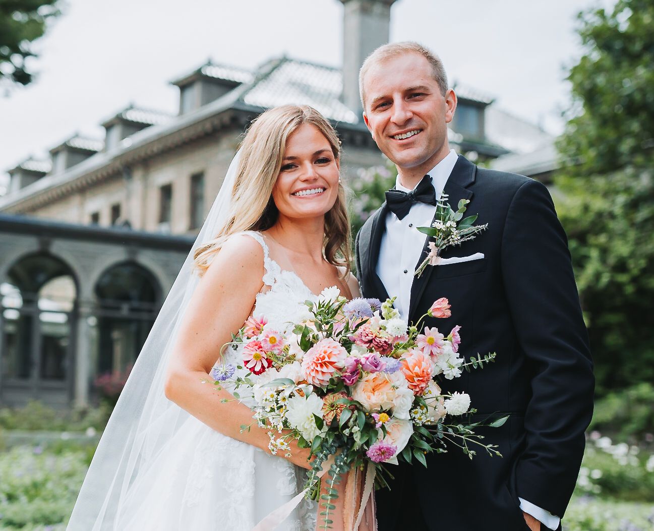 Wedding couple poses outdoors holding vibrant floral bouquet near white stone building.