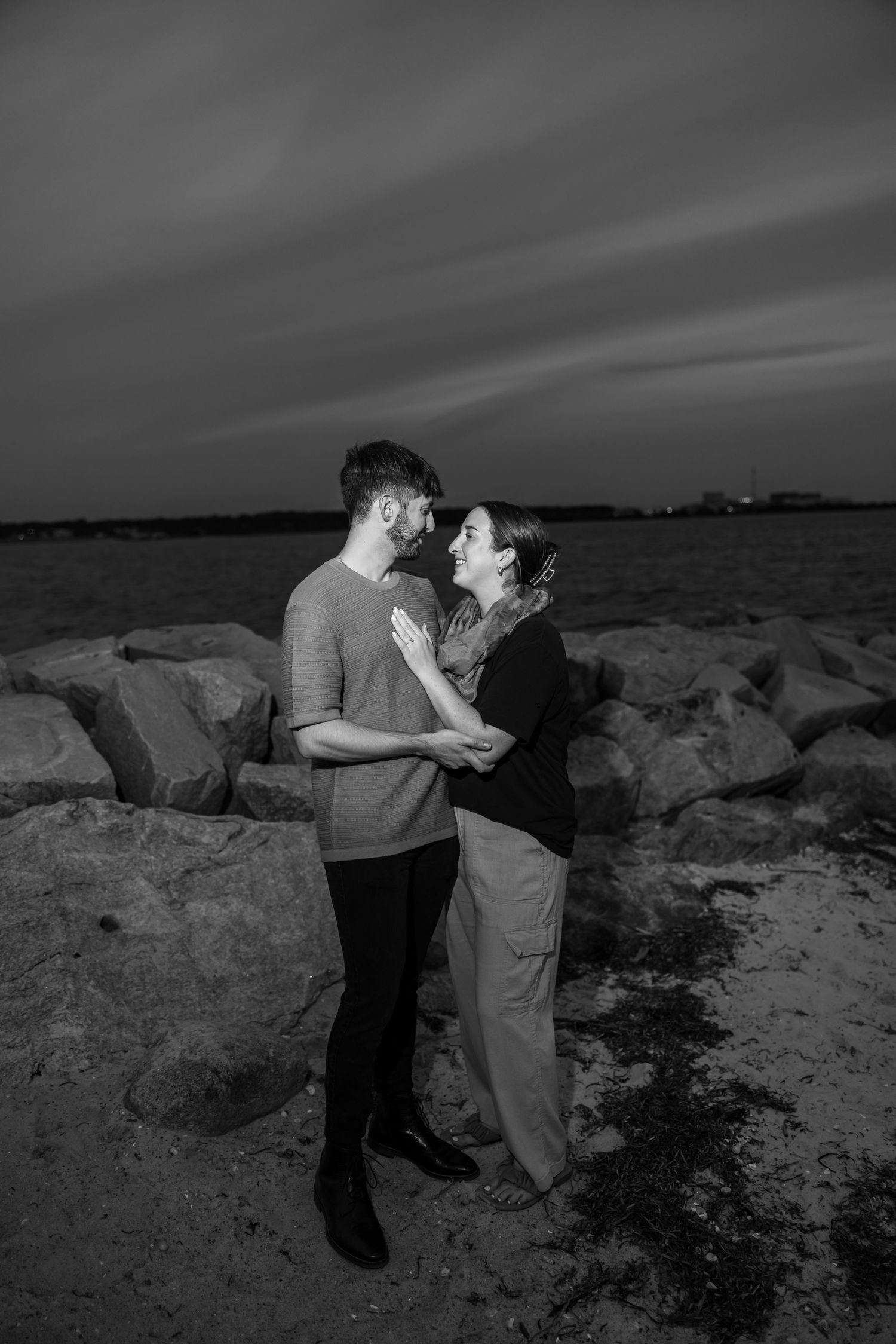A dramatic black and white seaside engagement photo series on rocky coastal terrain.