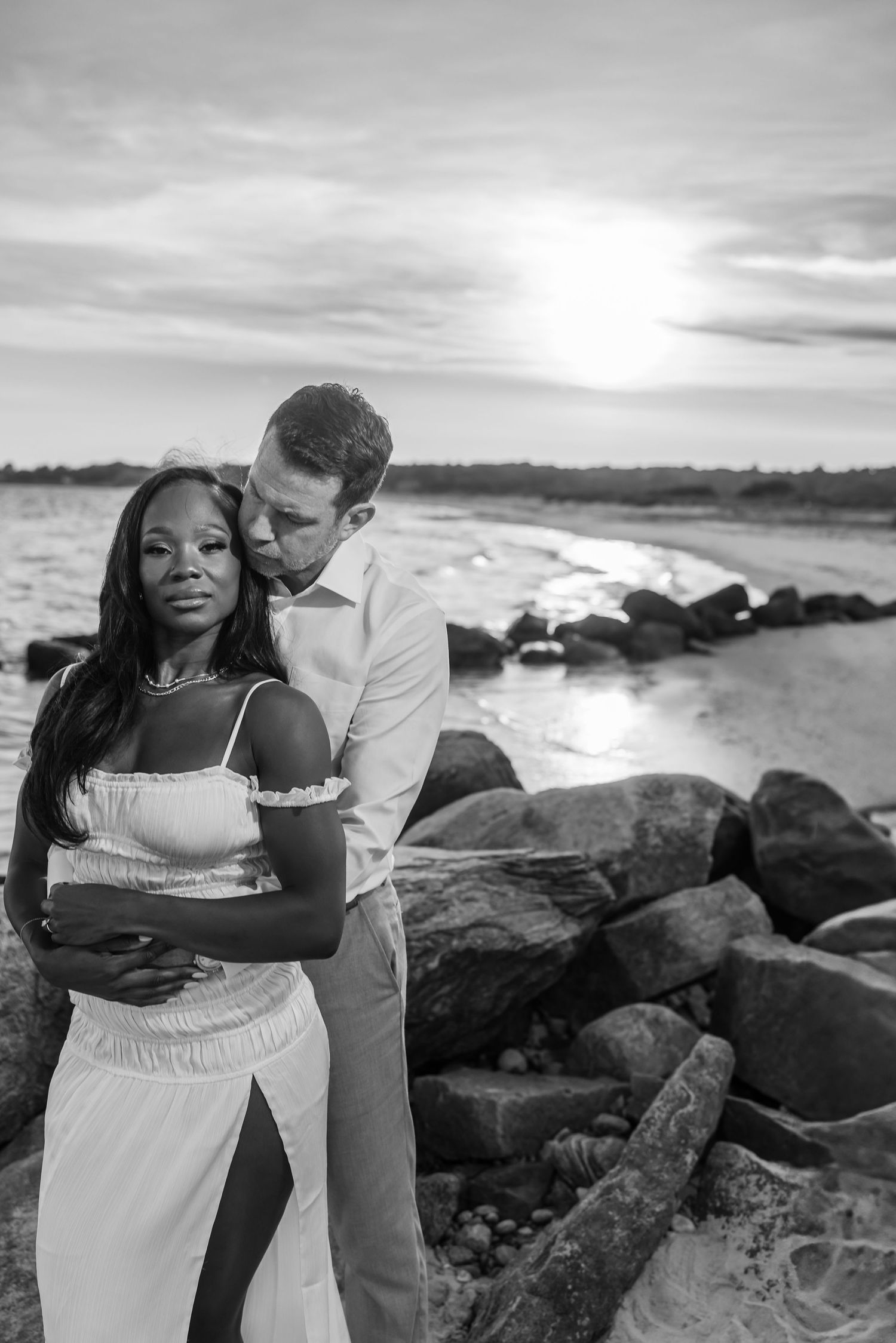 Black and white beach photo of a couple embracing. One person wears white and stands in front while their partner in light clothing embraces