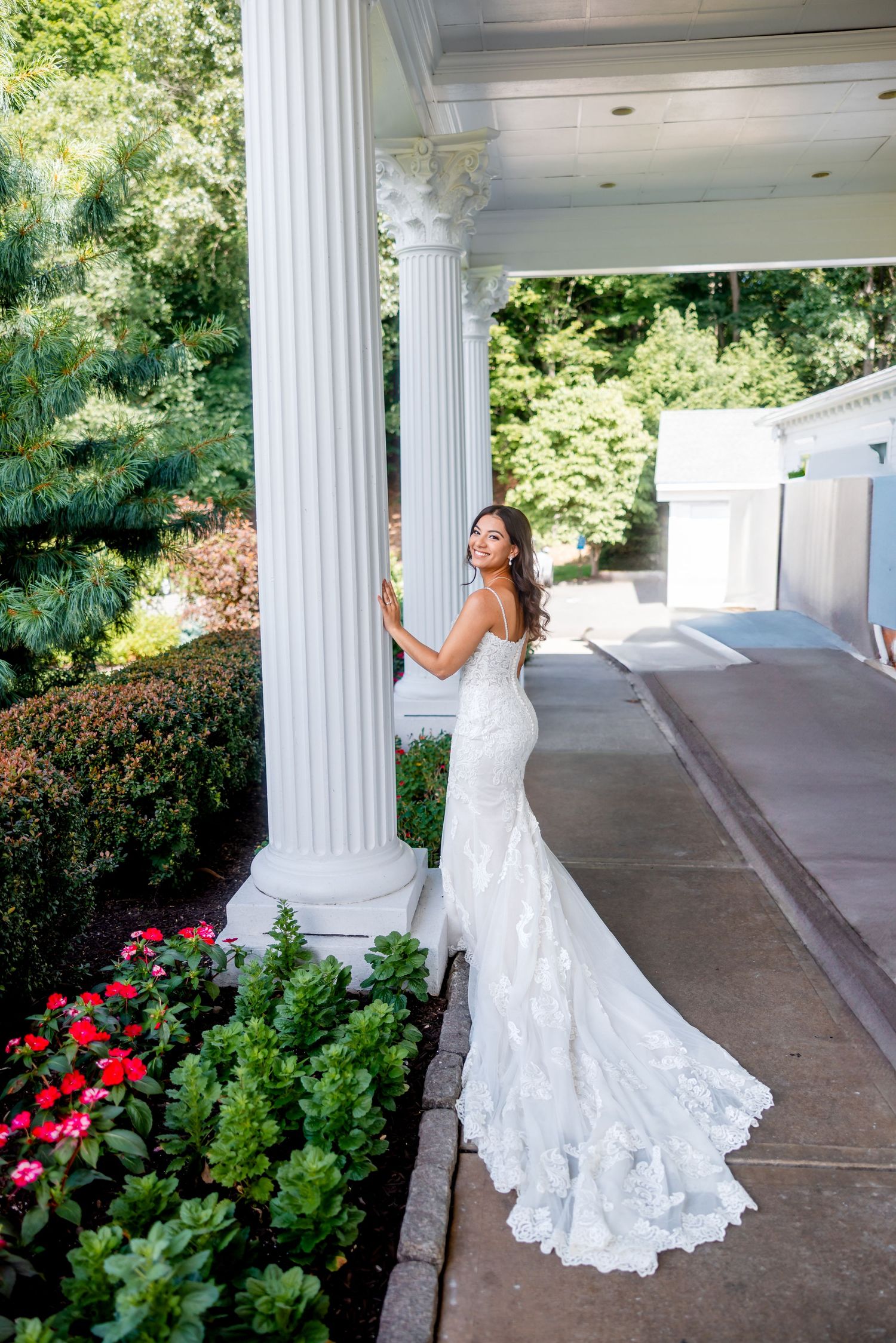 Bride in a fitted lace wedding gown with long train poses by white classical columns and garden flowers.