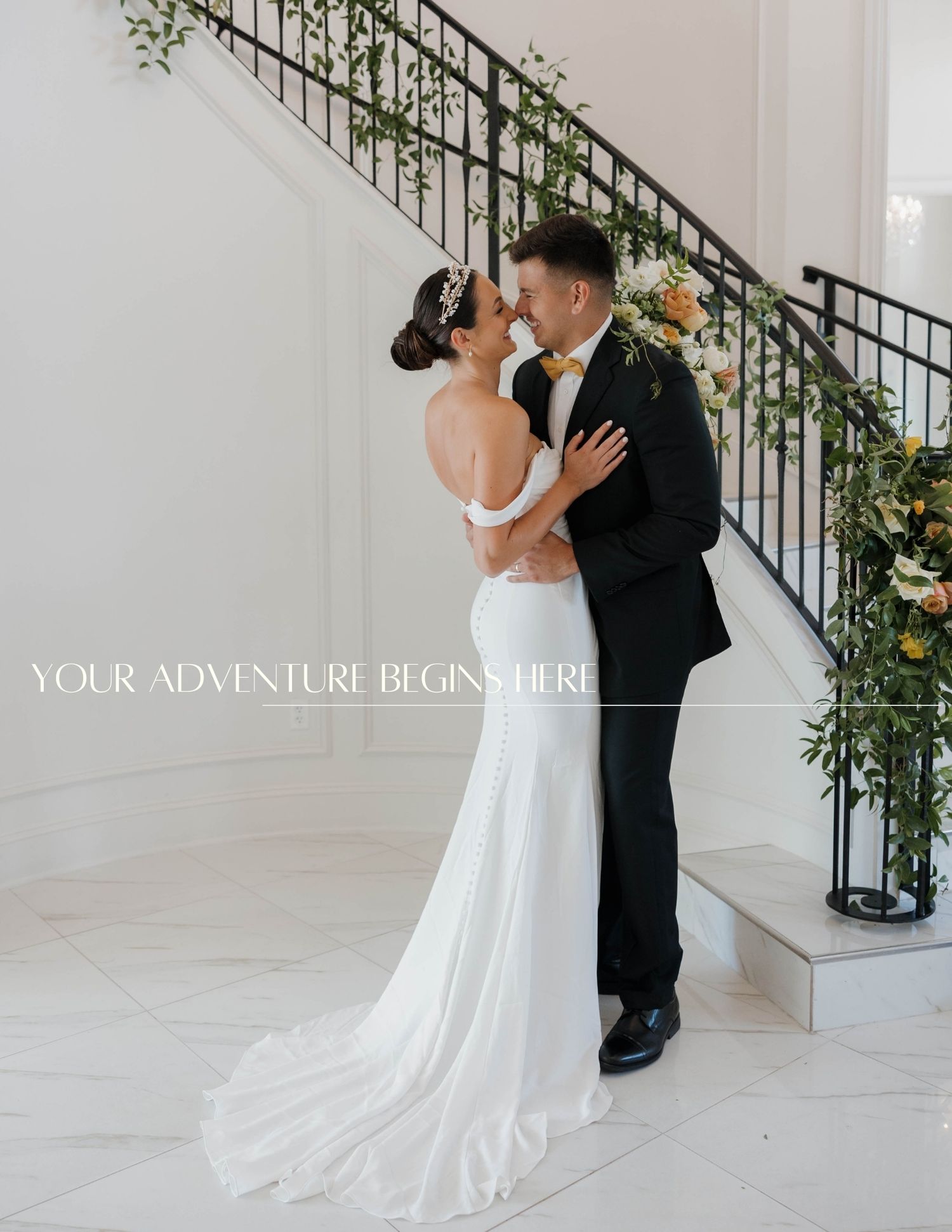 A romantic wedding couple embraces on elegant white stairs decorated with floral greenery and text overlay.