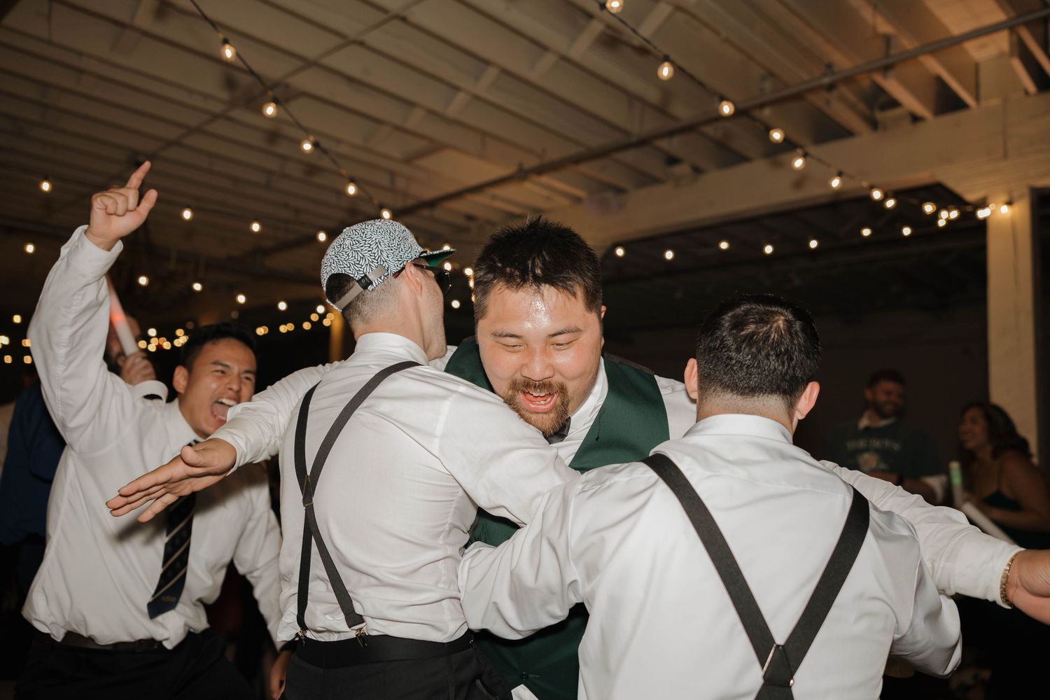 Groomsmen in white shirts and suspenders dancing and celebrating at a wedding reception with string lights overhead.