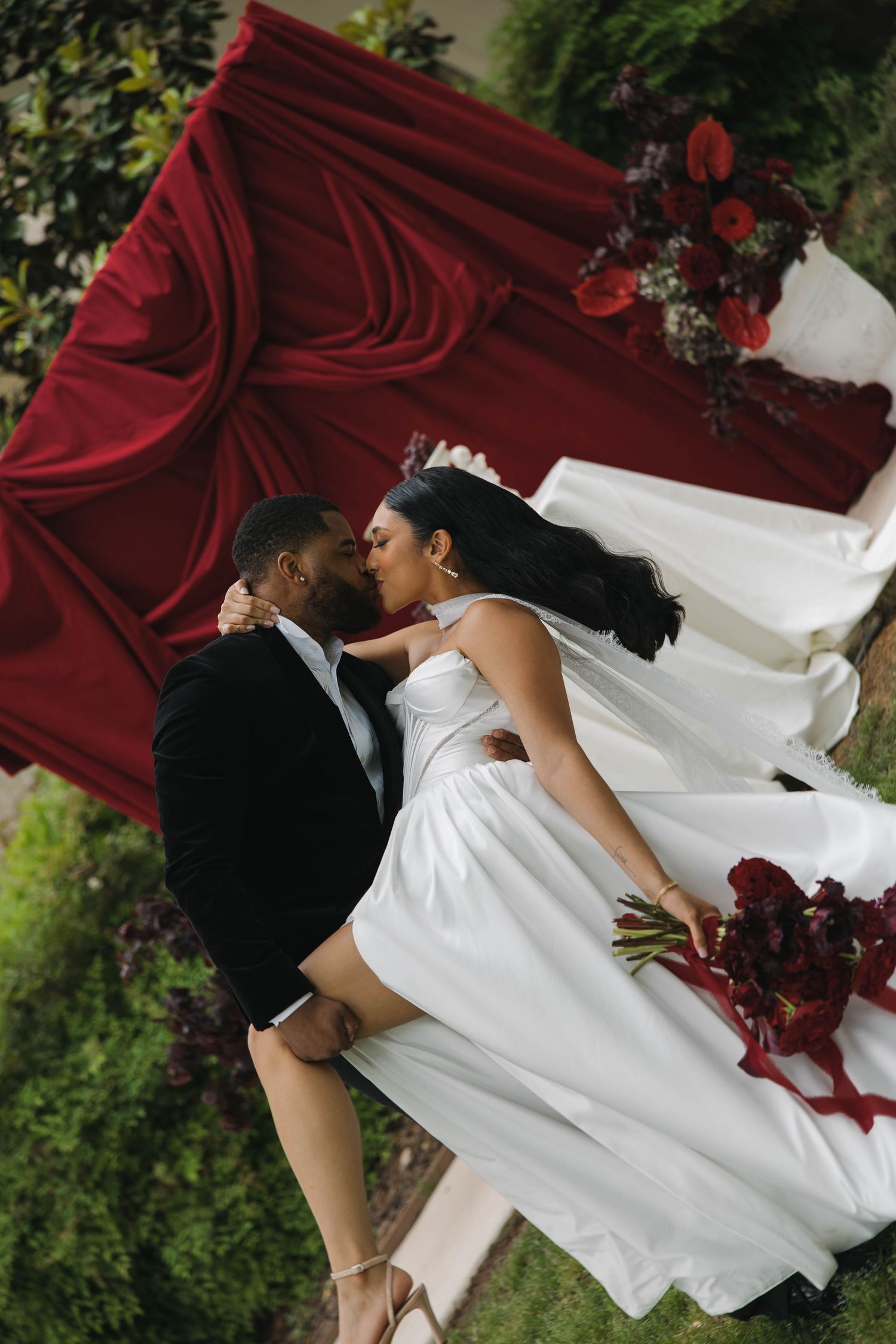Newlywed couple sharing an intimate moment on red fabric with bride in flowing white dress and burgundy floral details.