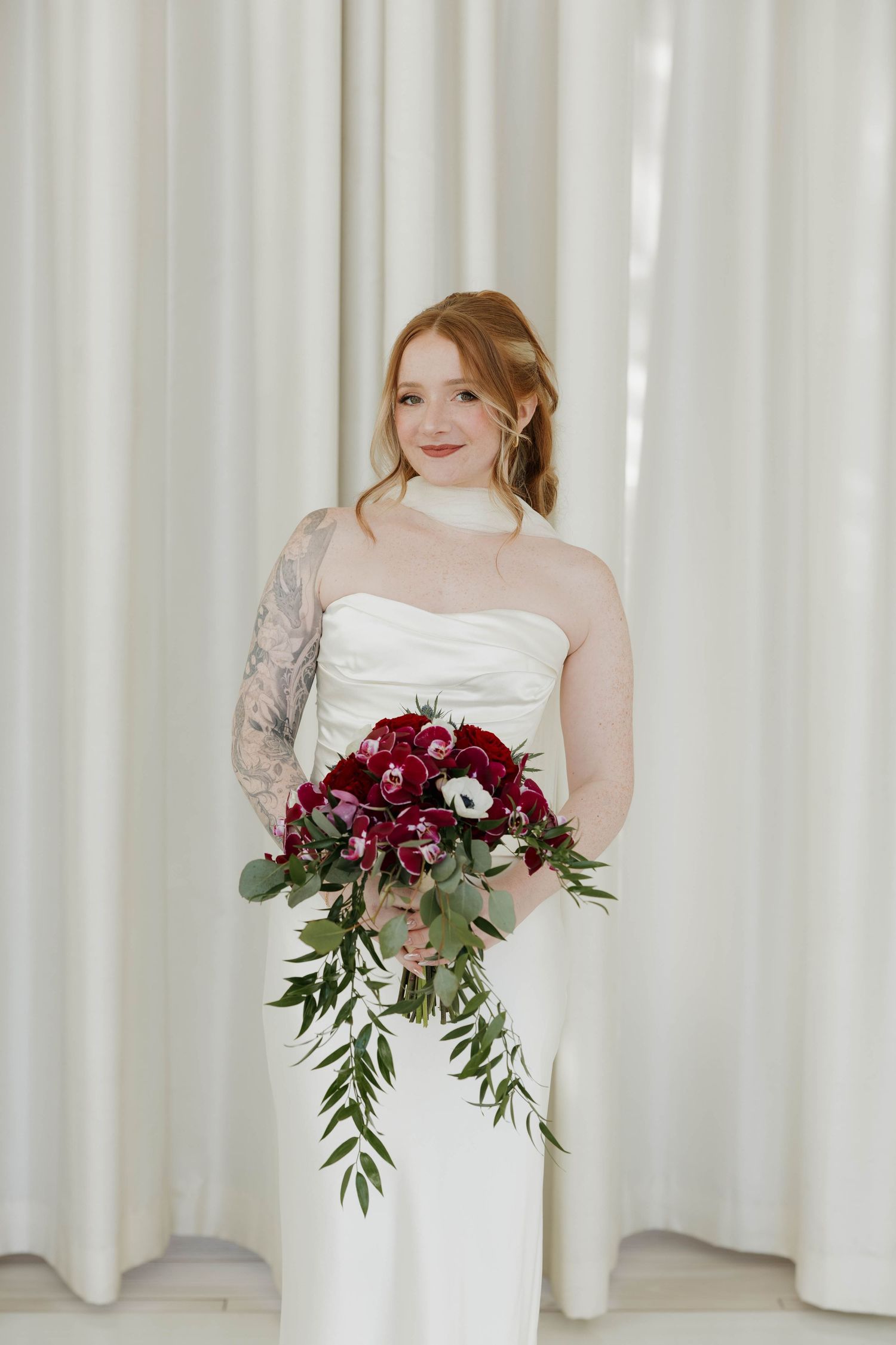 Bridal portrait showing a white wedding dress and deep burgundy floral bouquet against white curtain backdrop.