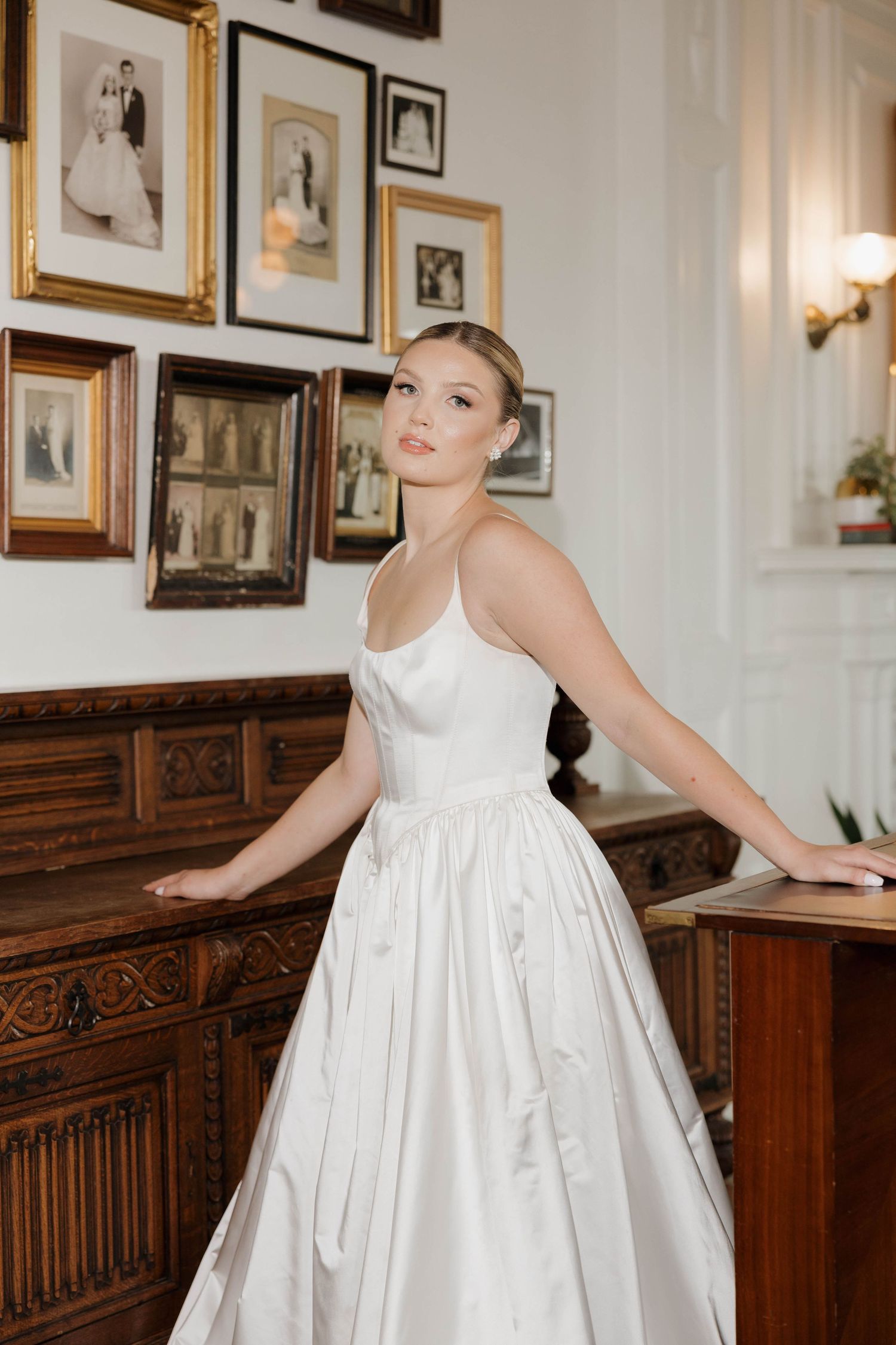 A person in a flowing white dress poses elegantly against a wall decorated with framed photographs and wooden paneling.