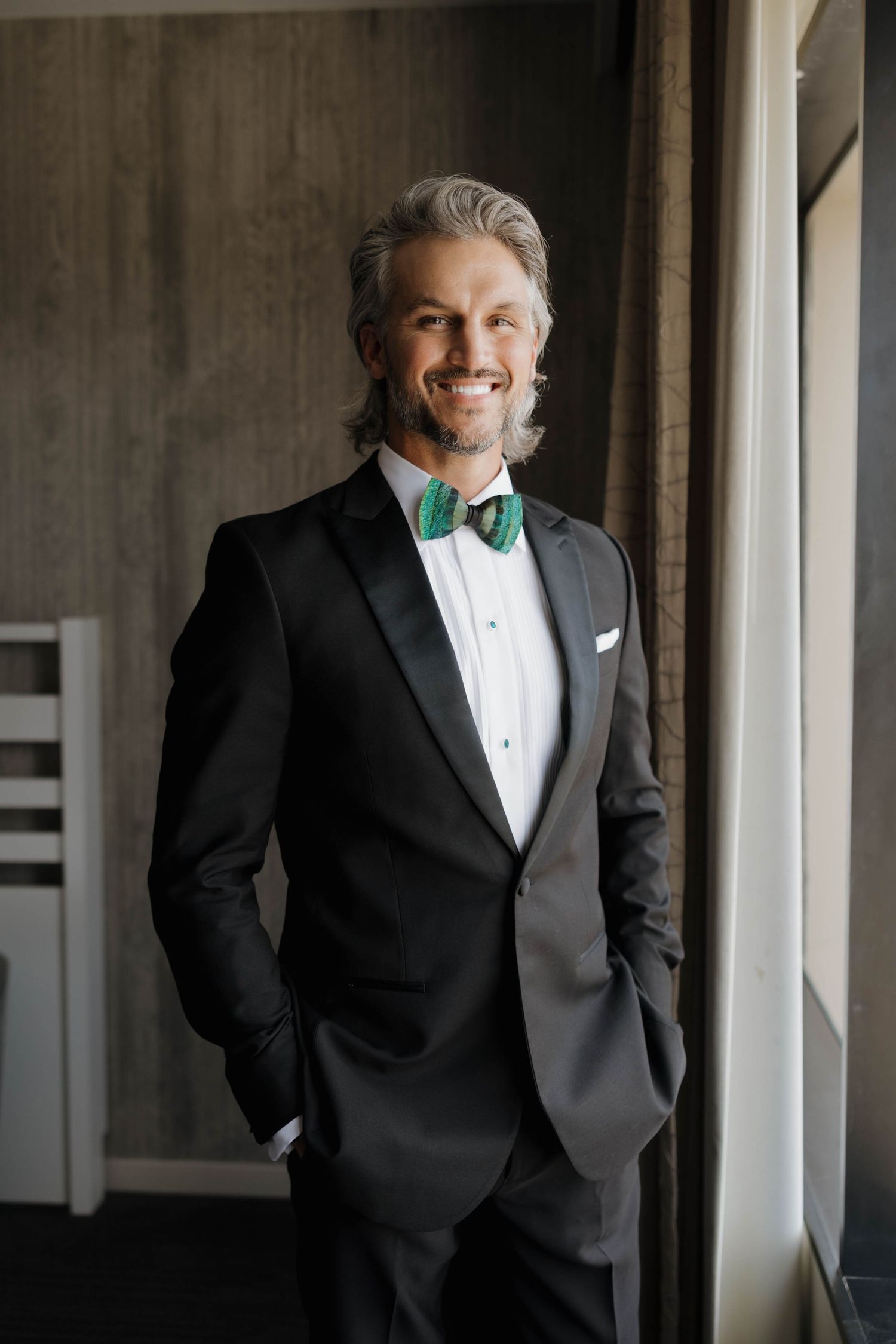 Groom in classic black tuxedo and green bow tie poses by window in formal portrait.