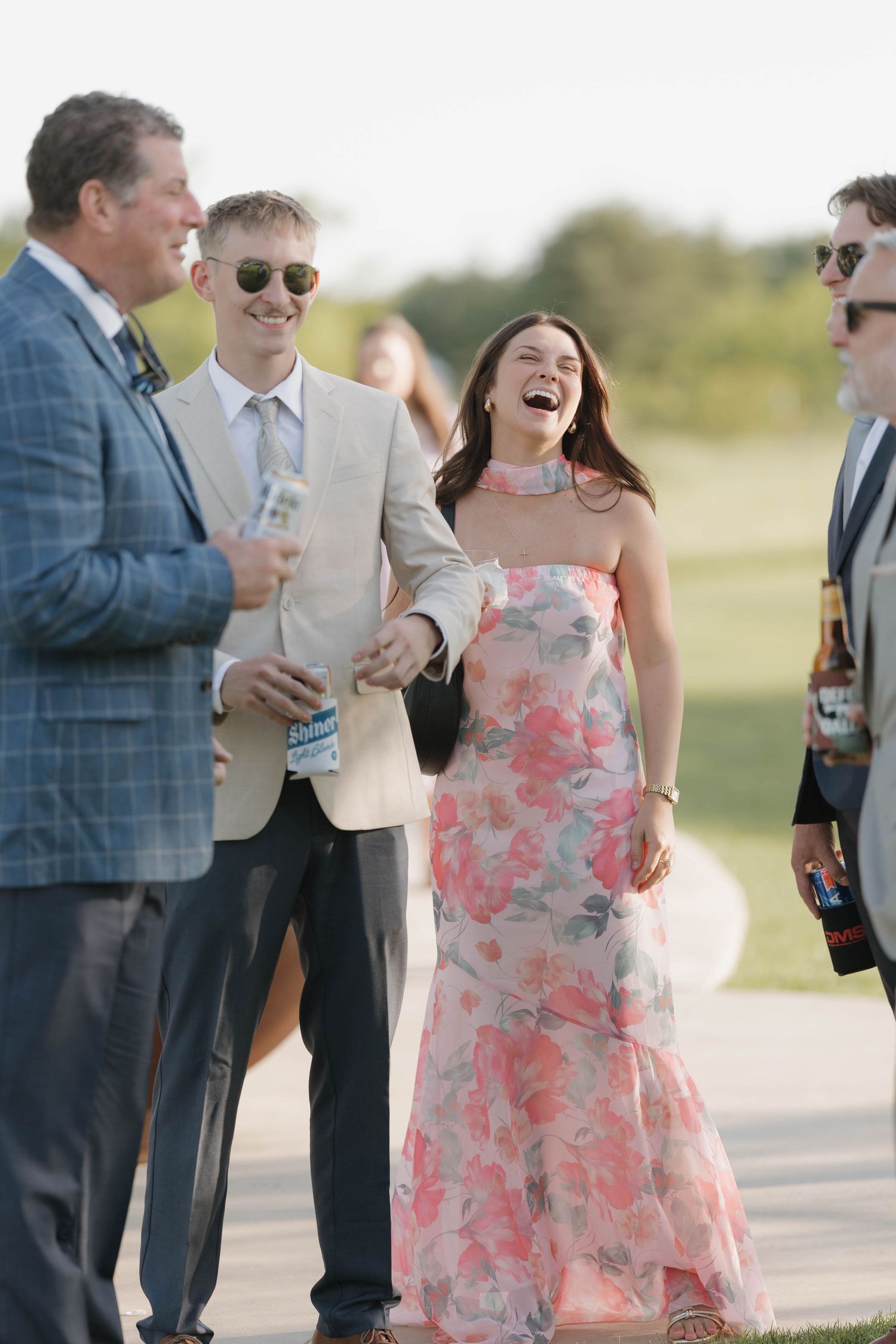 A group of well-dressed guests share a lighthearted moment at an outdoor event with someone in a pink floral gown.