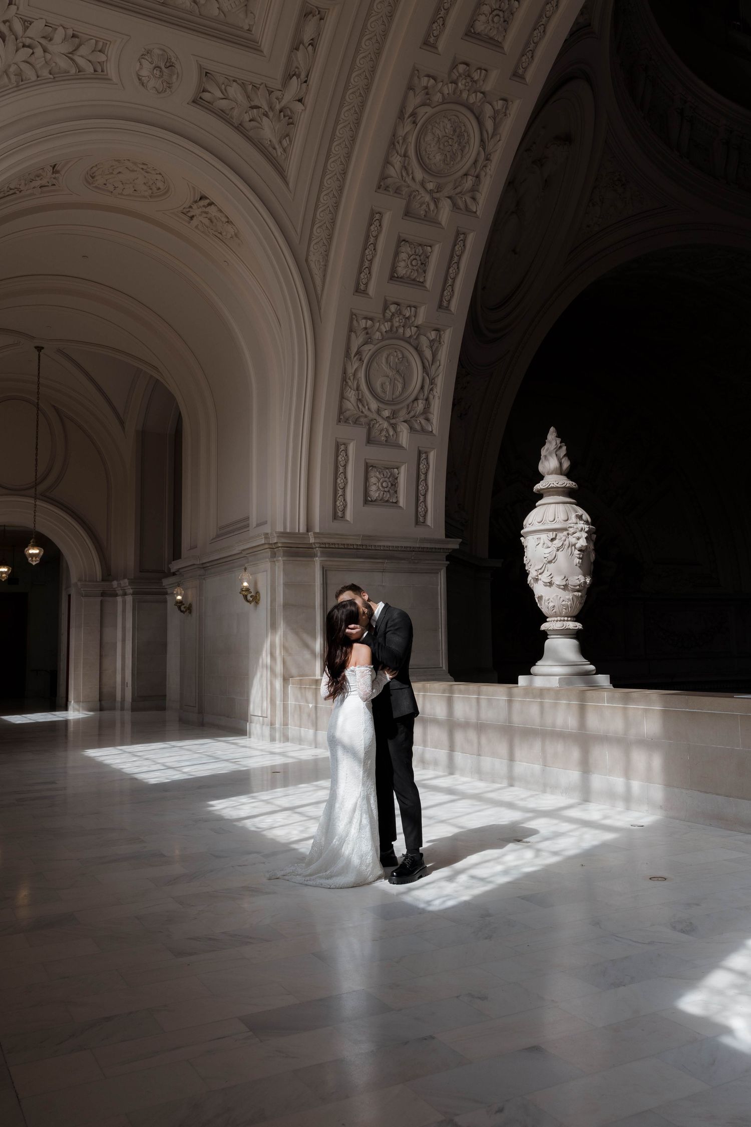 A bridal couple stands in a grand white baroque hallway with elegant architectural details as sunlight streams through windows.