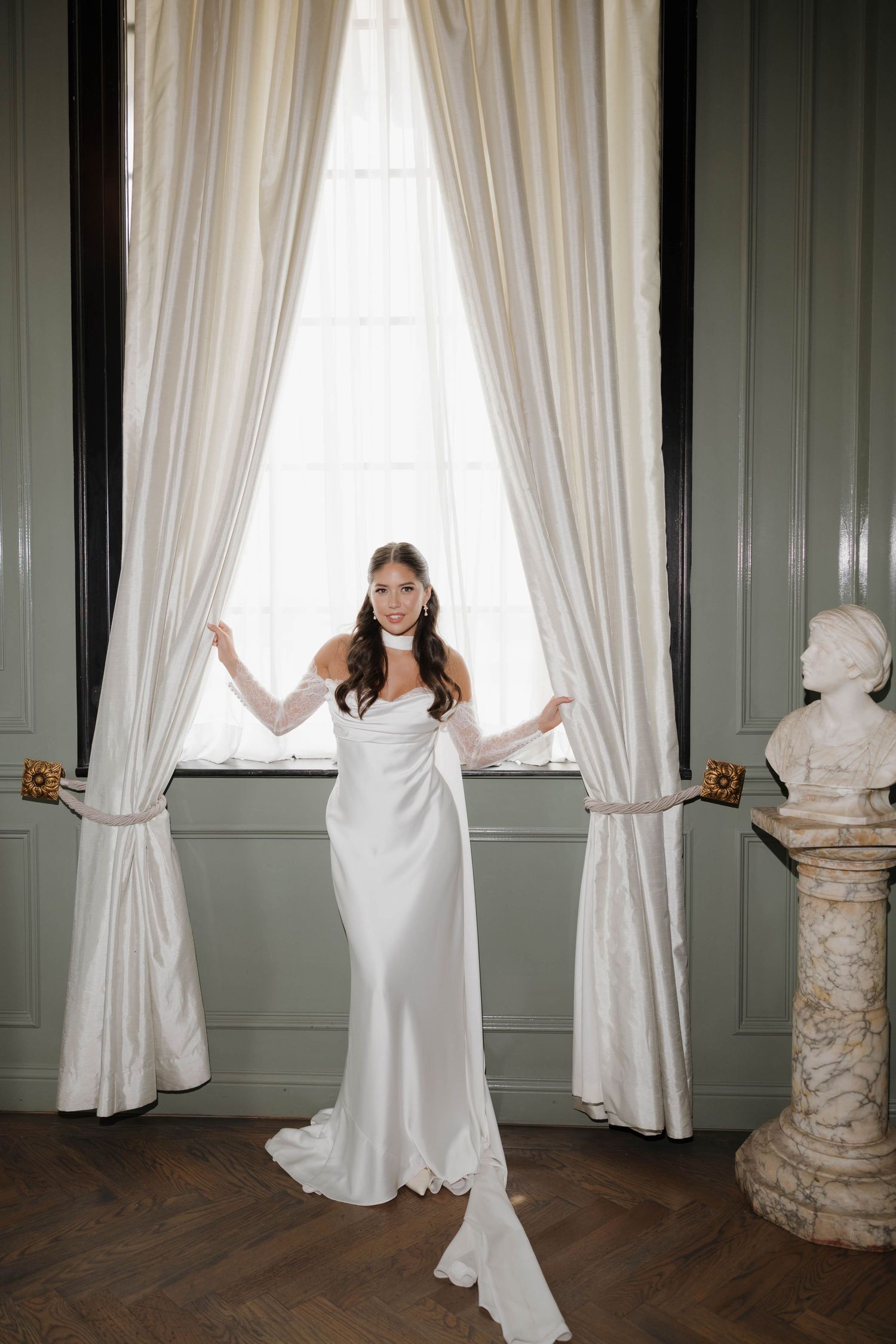 Elegant bride in flowing white silk gown stands between cream curtains in classic room with ornate marble pedestal.