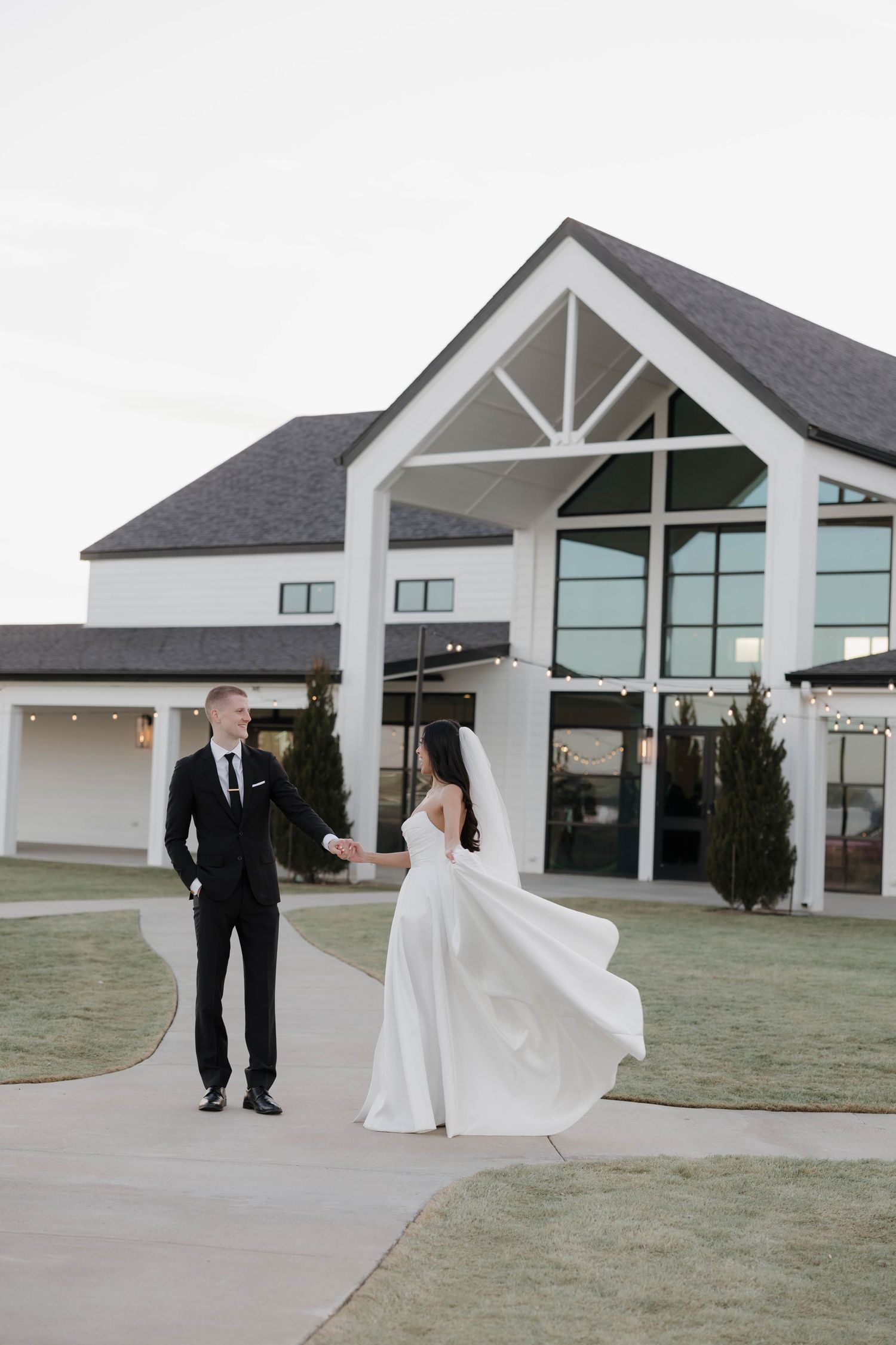 A couple in wedding attire share a moment outside a modern white church building with large windows.