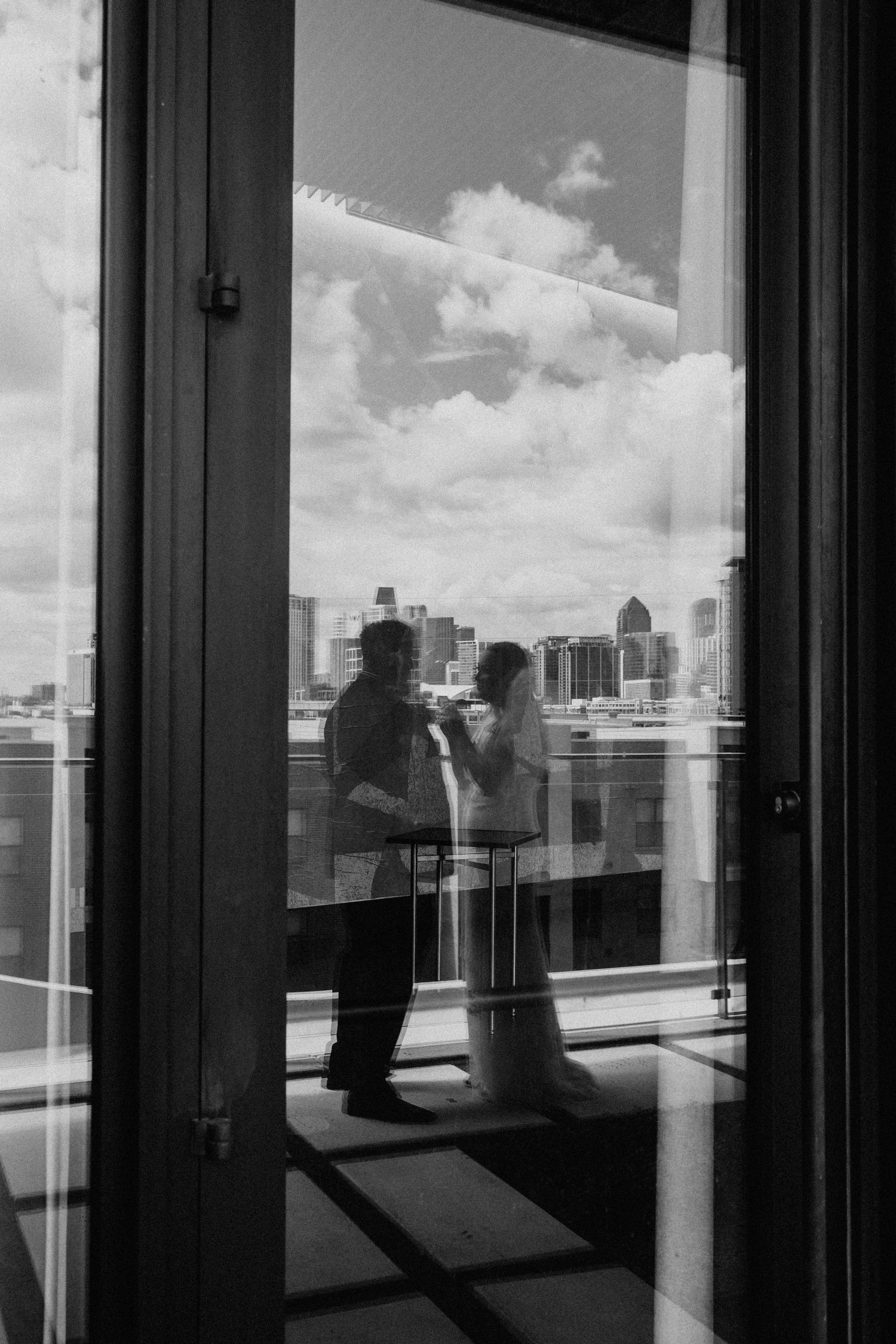 A black and white cityscape view through floor-to-ceiling windows with reflections and silhouettes against a cloudy sky.