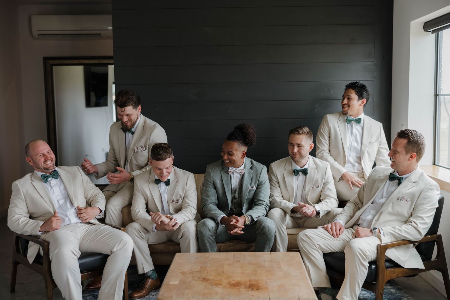 A group in light beige suits relaxes on a couch and chairs in a modern minimalist setting before a special event.