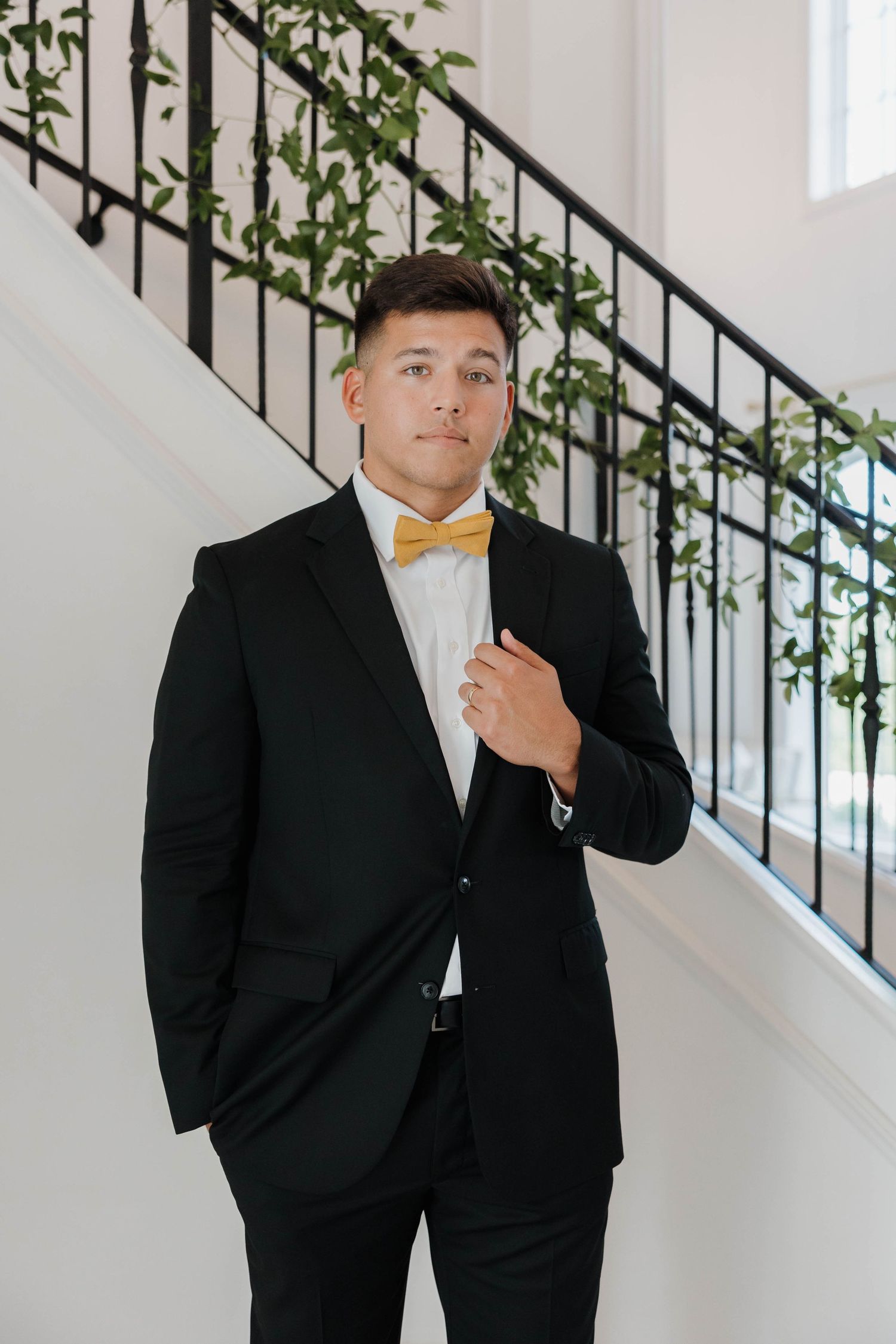 A person in a black suit poses on a white staircase with greenery and iron railings in an elegant indoor setting.