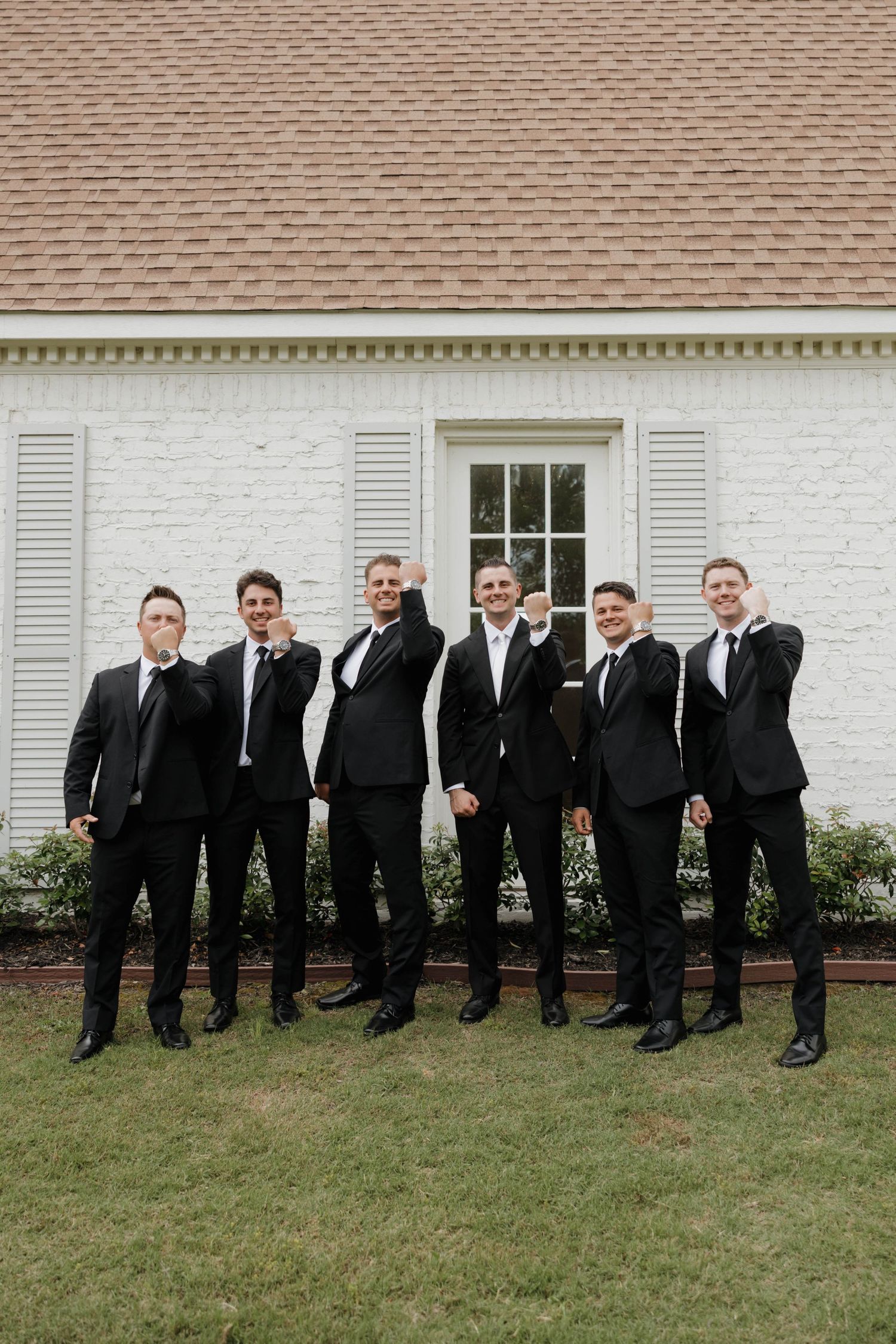 Wedding party of groomsmen in black suits and ties pose together on grass in front of white building.