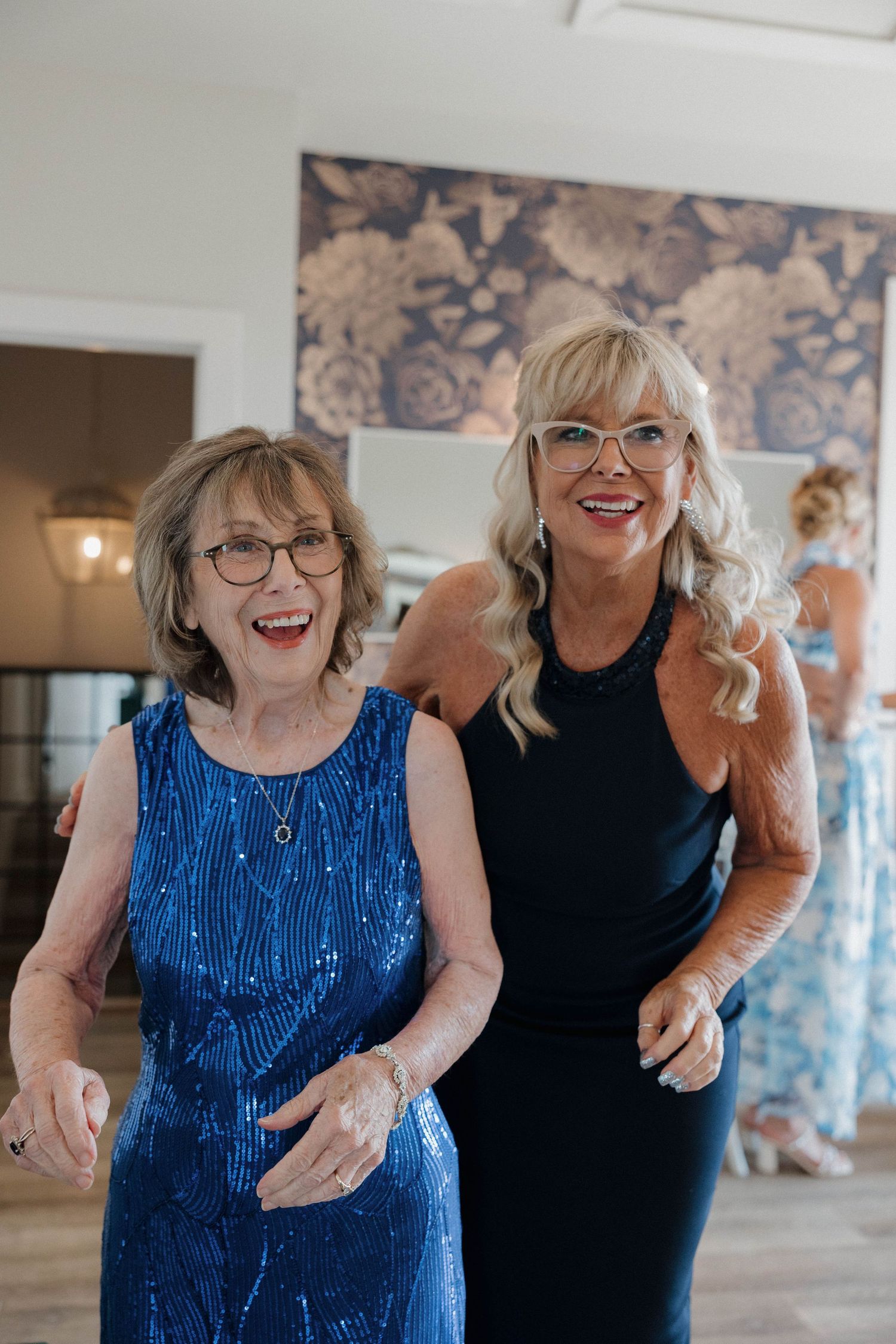 Two women in a joyful moment together, one wearing a sparkling blue dress and one in a black dress with decorative wallpaper behind.