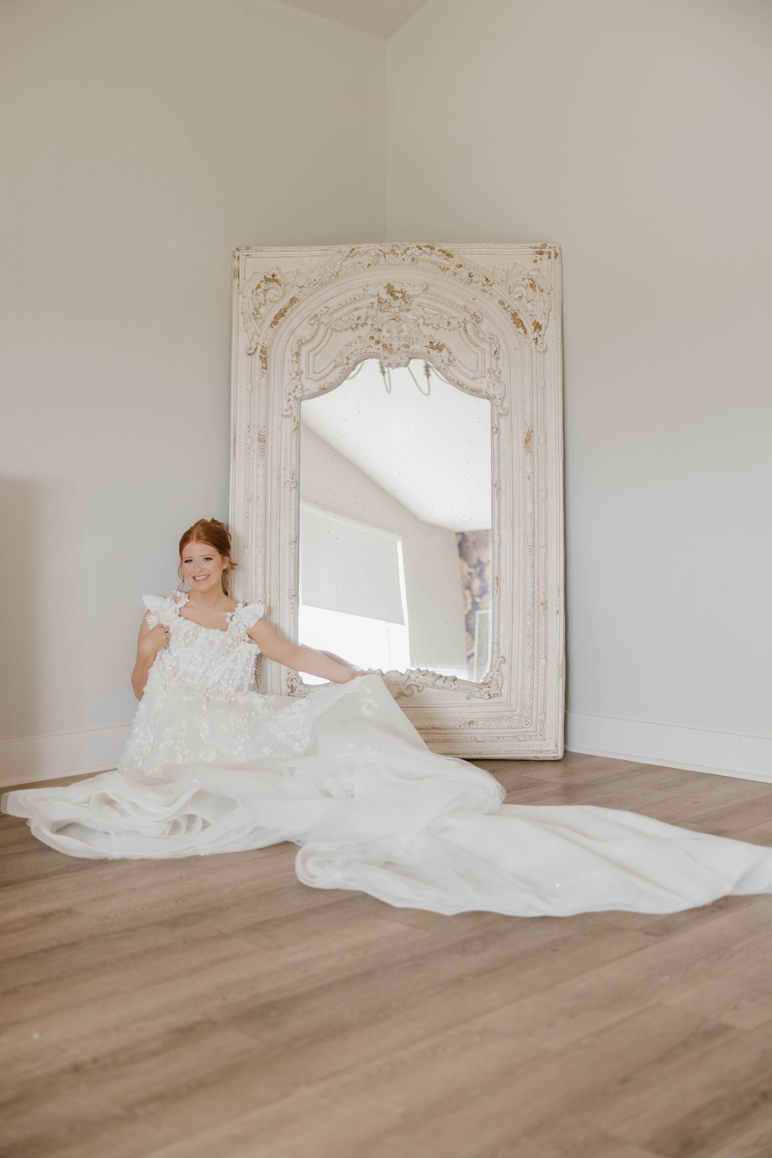 Elegant white wedding dress with long train spread across light wood floor beside ornate vintage white mirror.