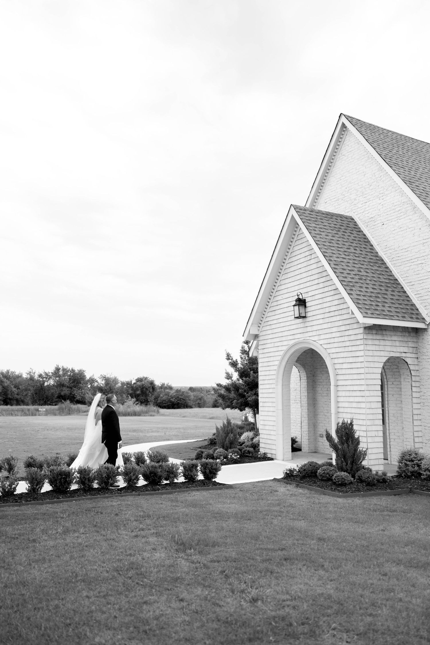 A black and white photo of a couple in formal attire standing outside a small white chapel with arched doorways.
