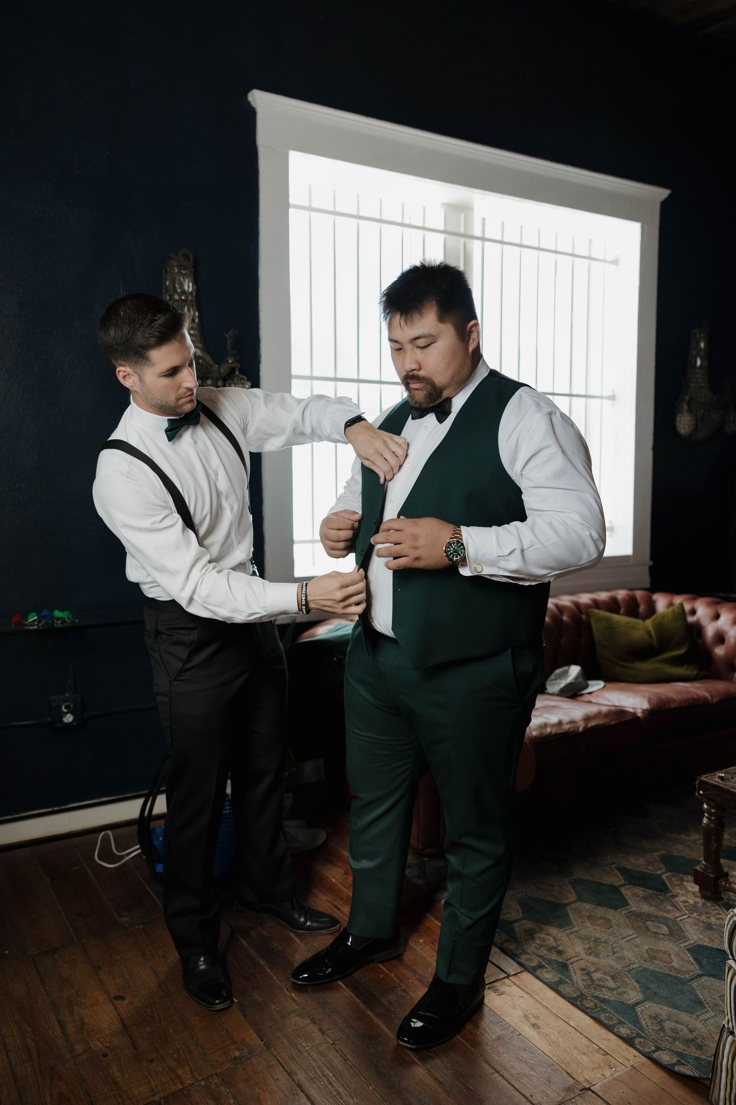 Two groomsmen in formal attire help each other get dressed in a dark room with hardwood floors and white trim.