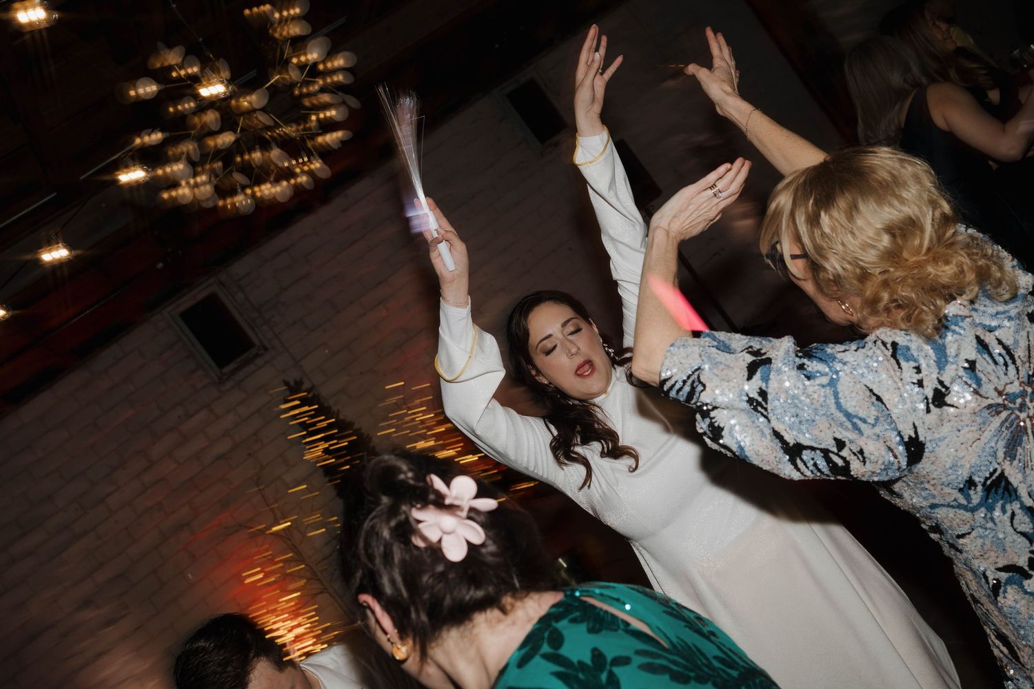 Wedding guests enthusiastically dancing with raised arms on a dark dance floor with ambient lighting.