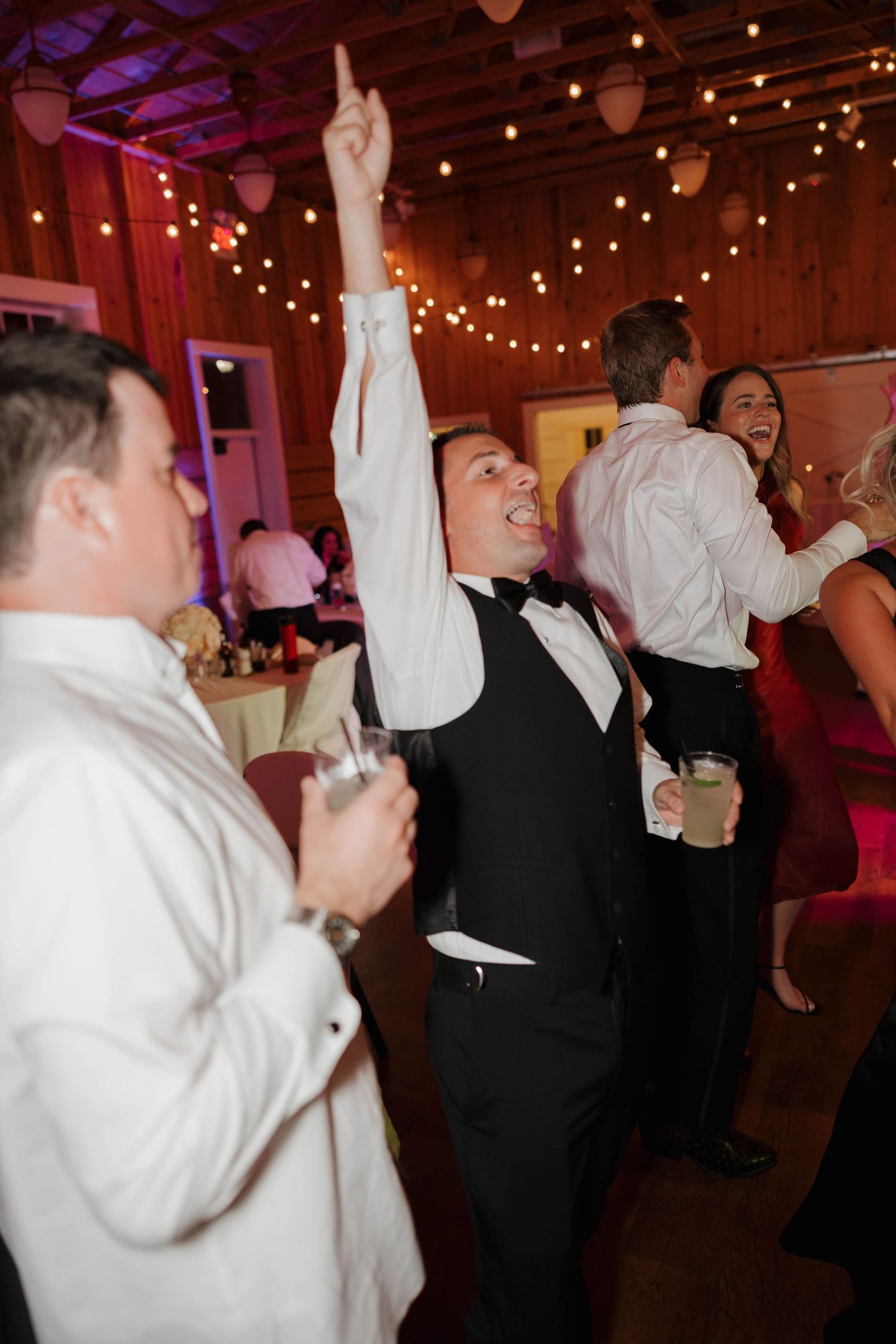 Party guests raise their arms in celebration on the dimly lit dance floor illuminated by warm string lights above.
