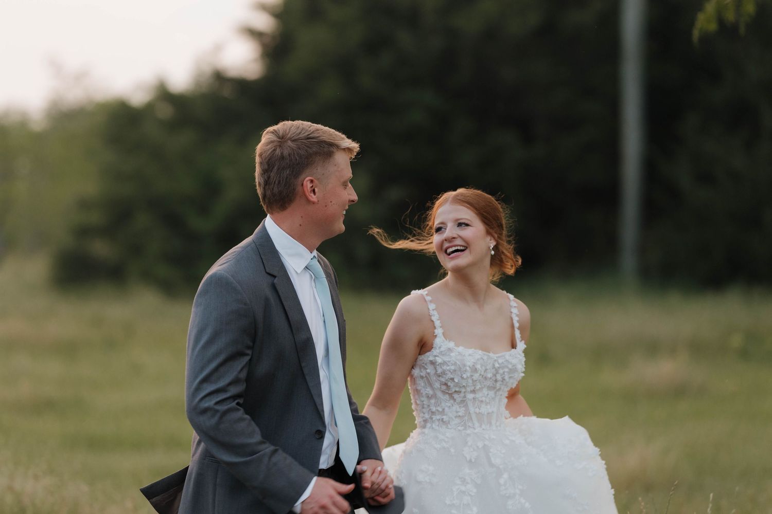 An elegant wedding couple shares a joyful moment together while standing in a serene outdoor field at sunset.