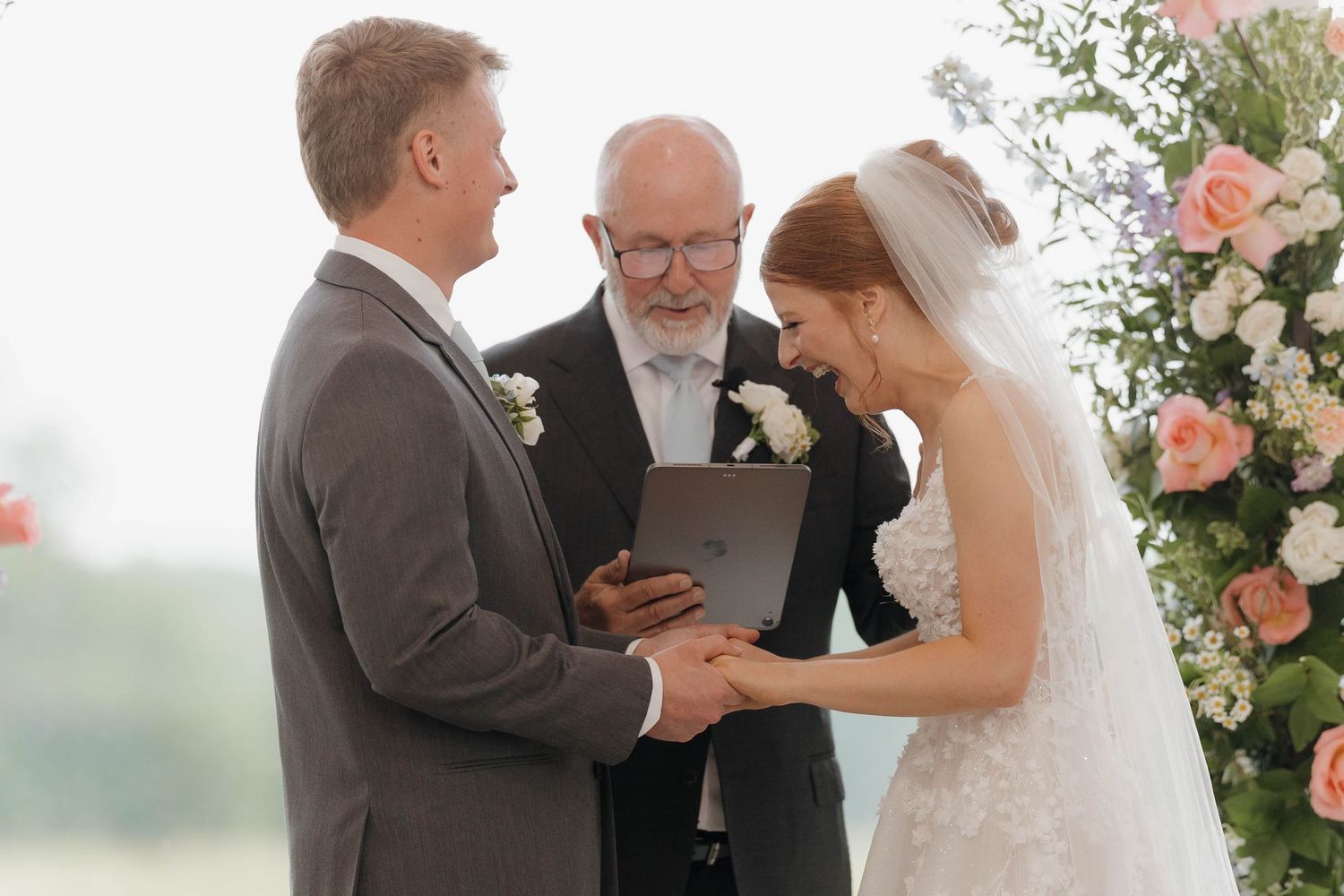 A couple exchanges vows at an outdoor wedding ceremony with pink floral arrangements and a suit-clad officiant.