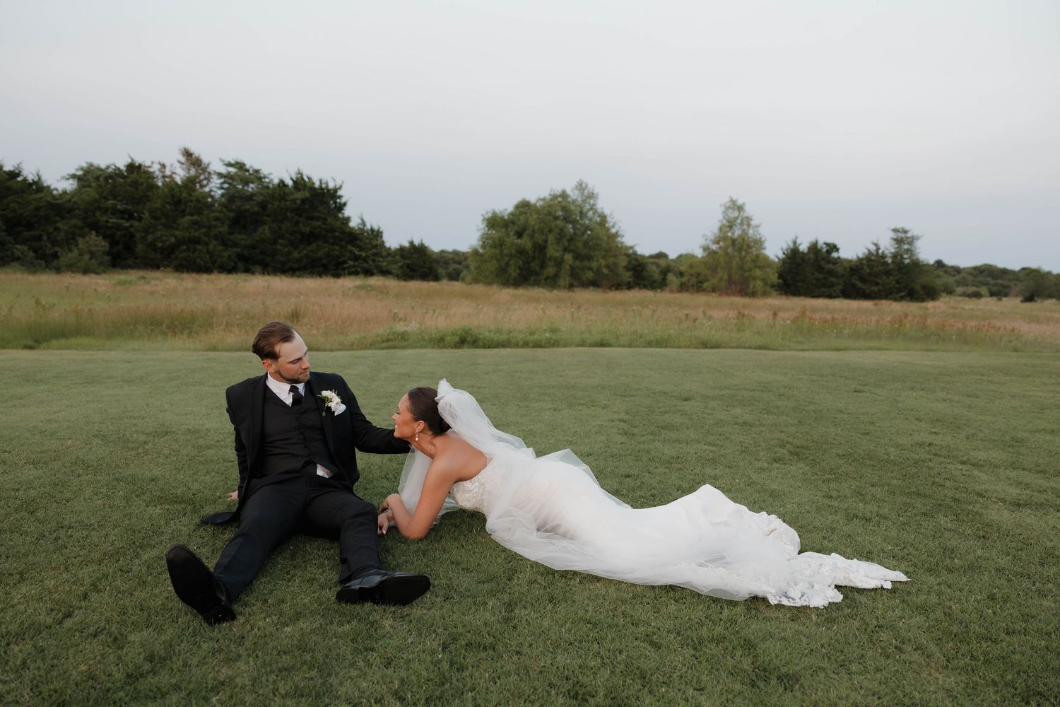 Couple in wedding attire laying in a green grassy field during an outdoor photo session at sunset.