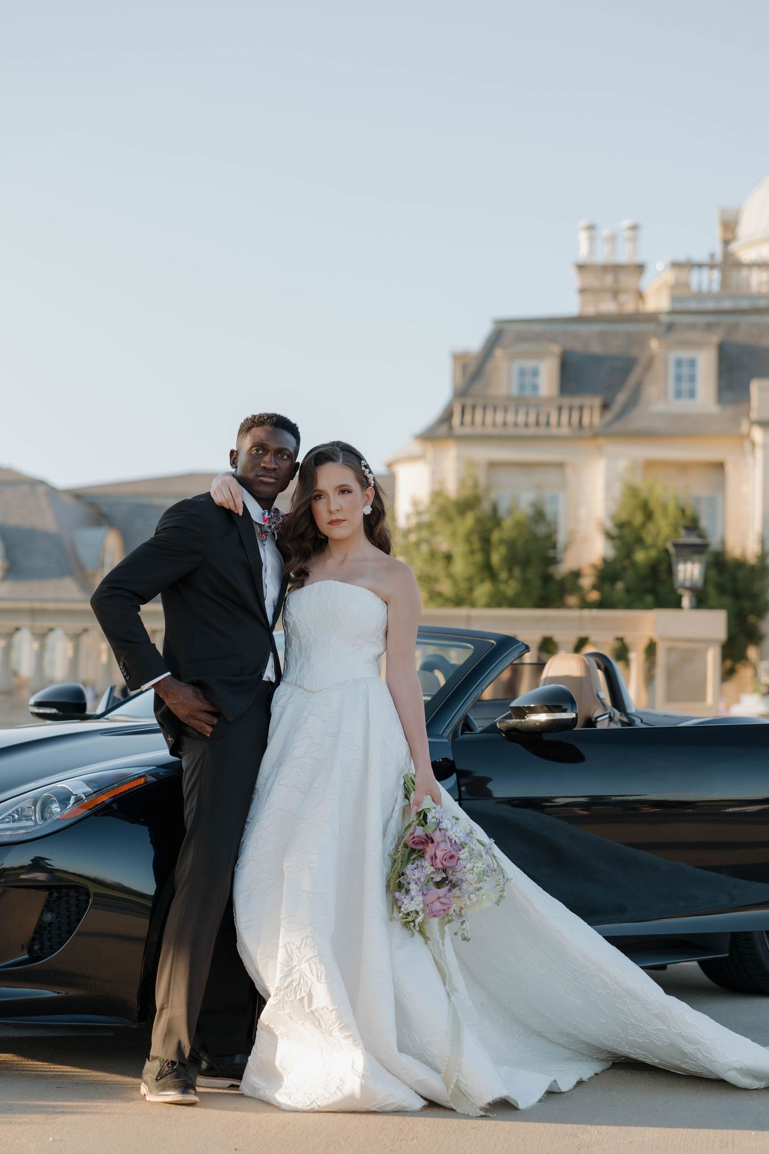 Elegant couple poses with vintage car at luxurious estate wedding venue during golden hour.