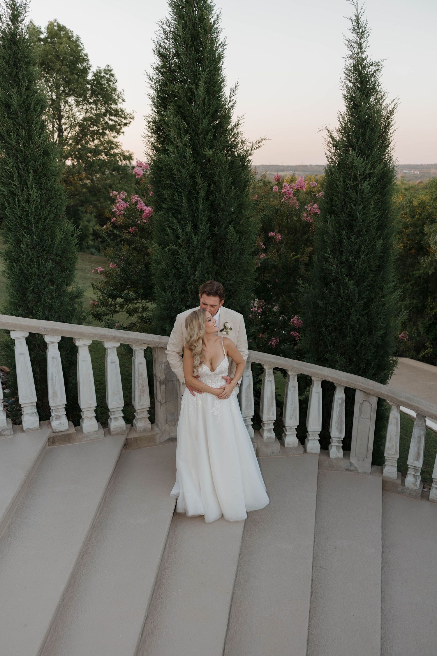 Couple sharing an intimate moment on a balcony with stone railings surrounded by evergreen trees at sunset.