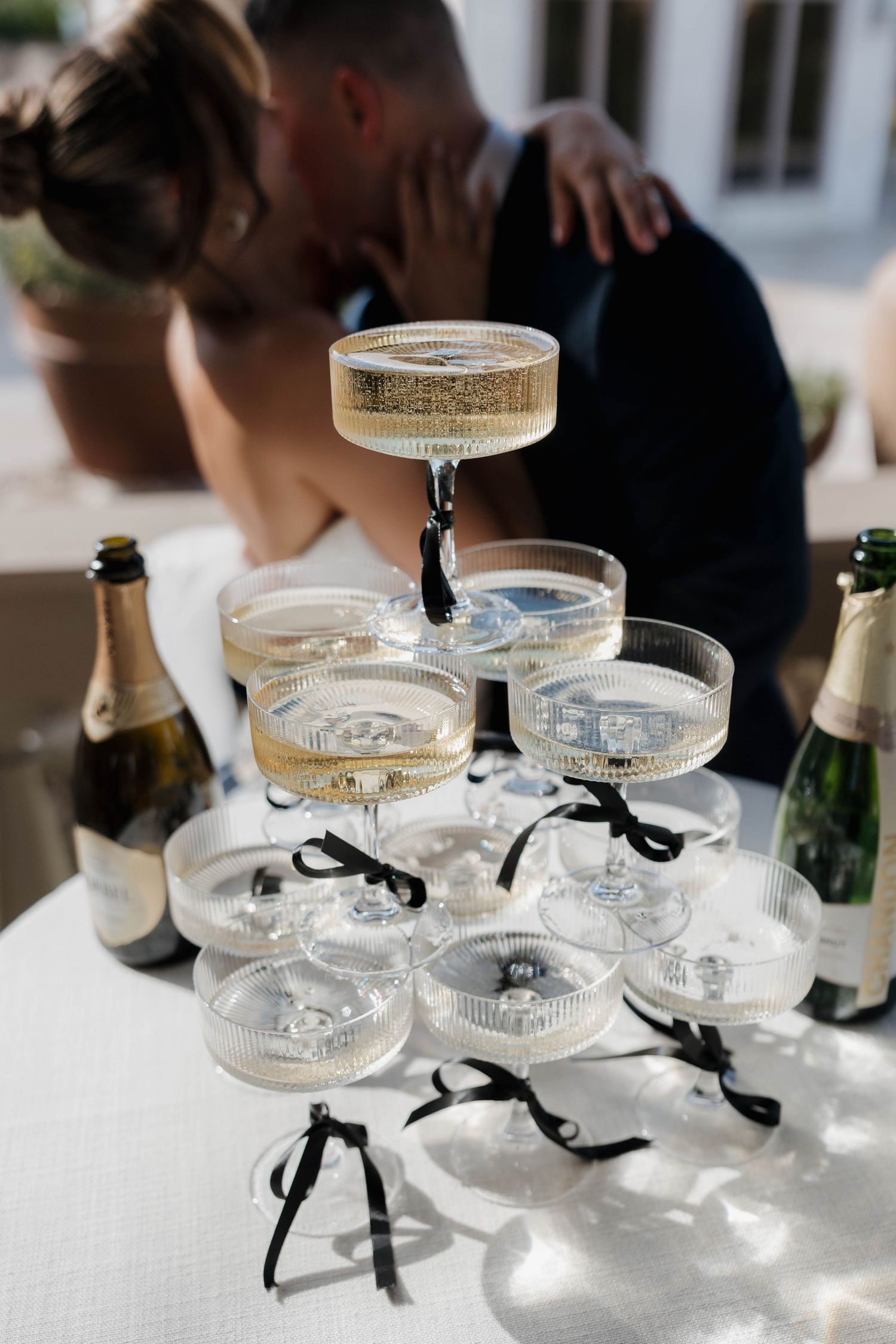 Champagne tower with black ribbons on coupe glasses arranged on white tablecloth with couple embracing in background.